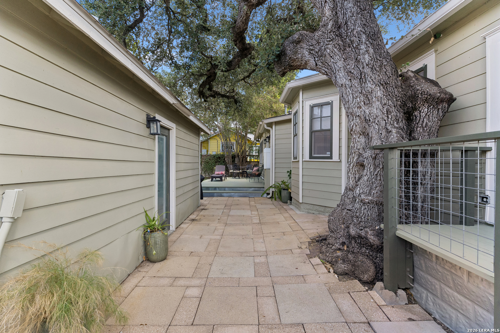 Paved courtyard between house wings, shaded by a massive mature tree with outdoor seating beyond.