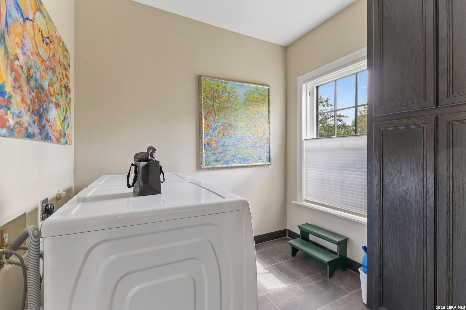 Laundry room with dark cabinetry, a sunny window, colorful artwork, and a green step stool.