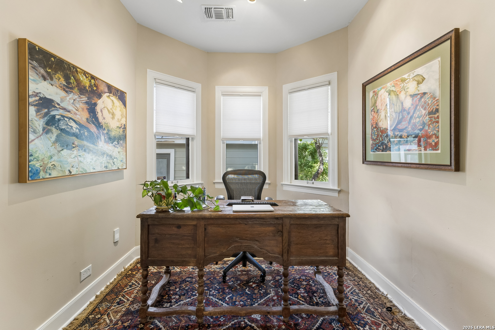 Home office with a wooden desk, three windows, framed artwork, and a patterned rug.