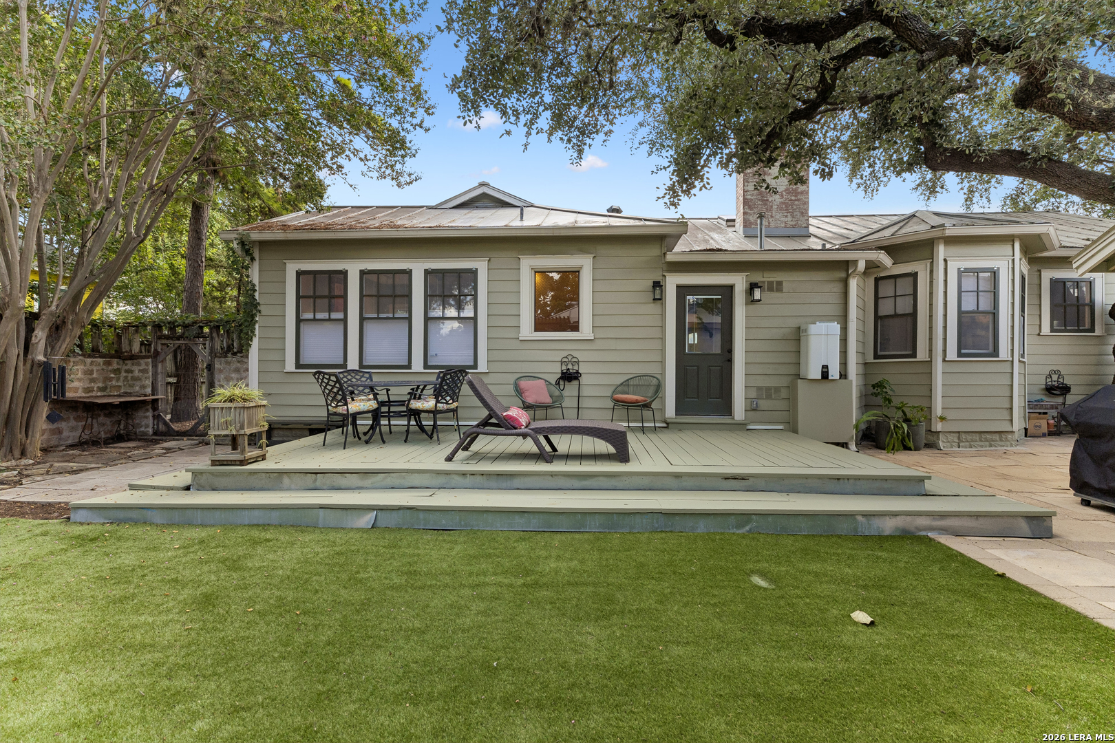 Rear of a single-story home with a furnished deck beneath mature shade trees.