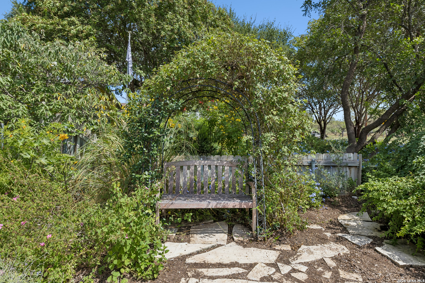 Garden bench beneath a leafy arbor beside a winding stone path and flowering plants.