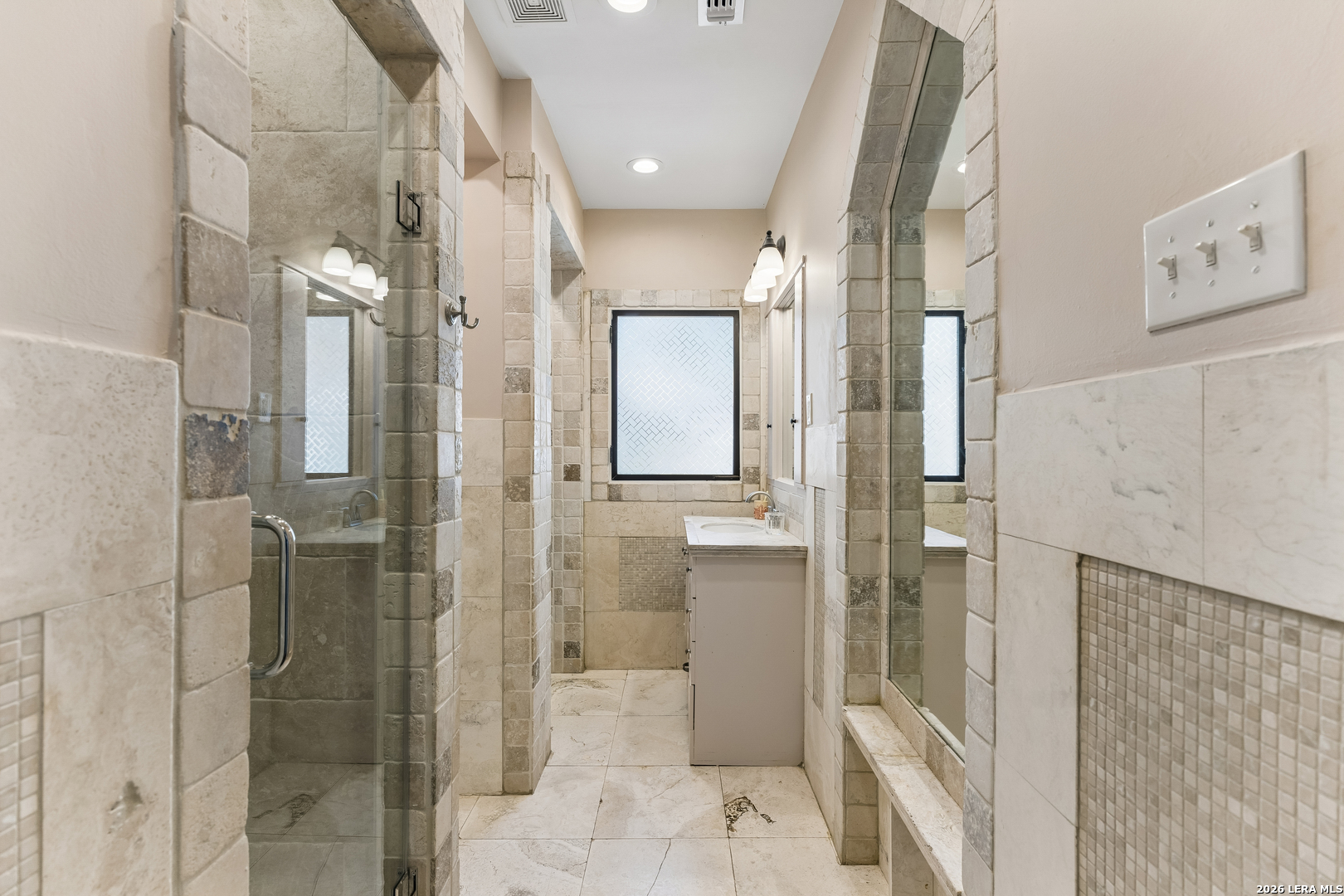 Bathroom with a glass shower, beige stone tile, white vanity, and large wall mirror.