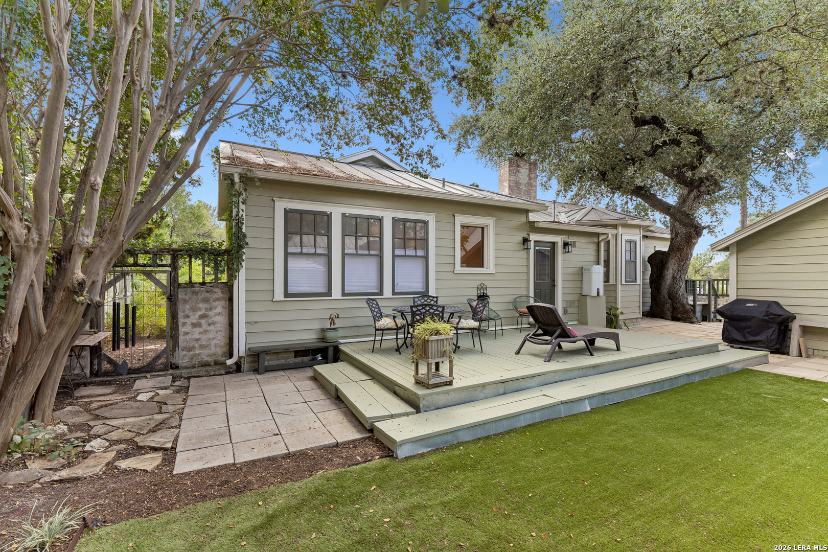 Home’s rear patio with a raised deck, outdoor seating, mature trees, and lawn.