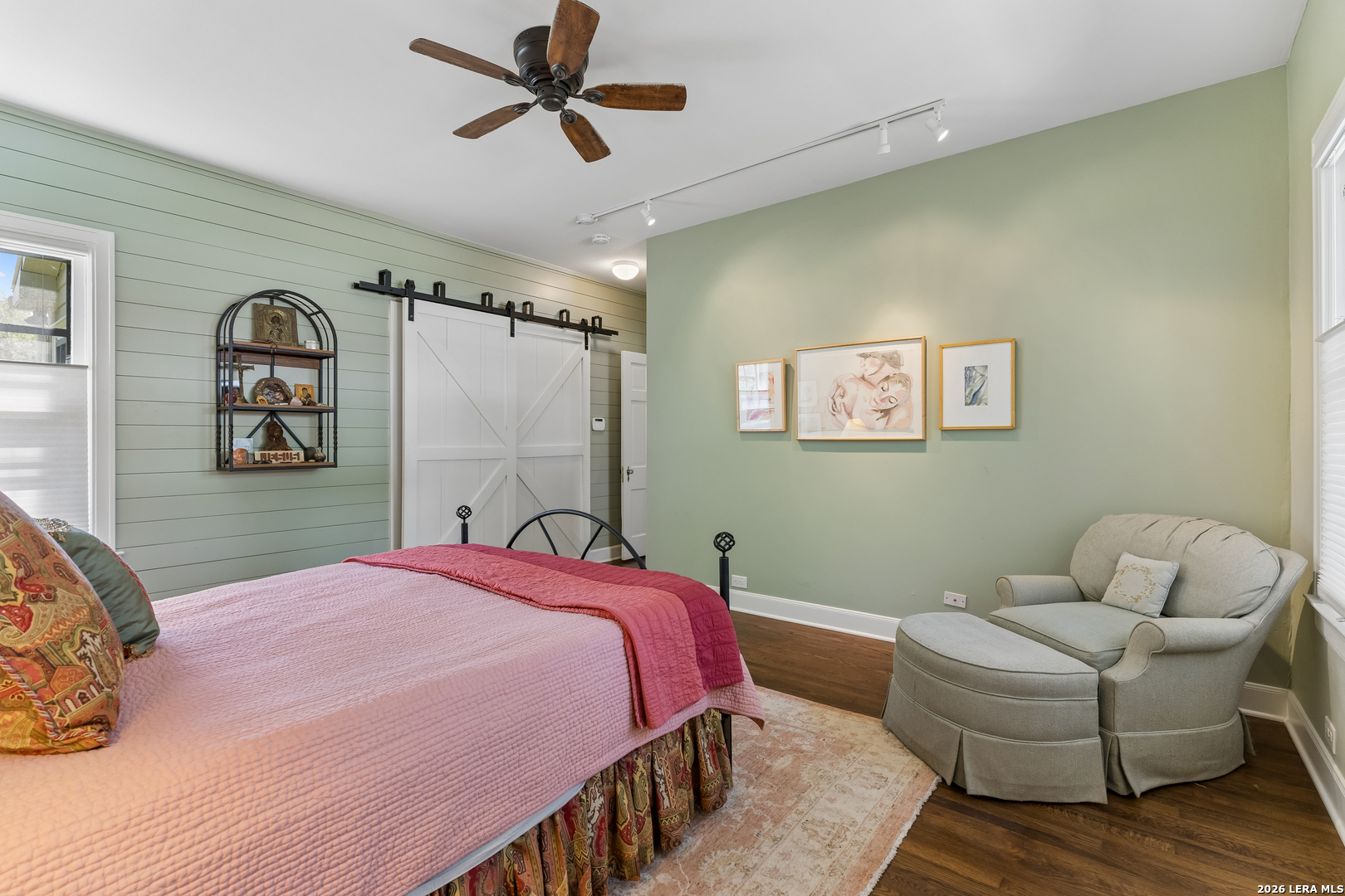 Staged bedroom with a pink bed, sage-green walls, sliding barn doors, and a gray armchair with ottoman.