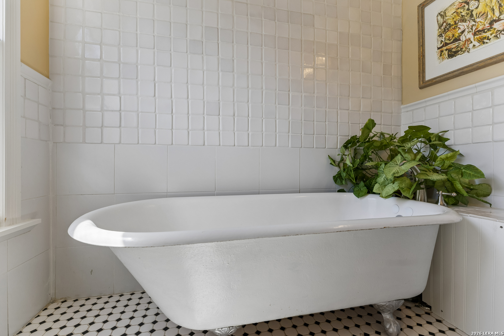 White clawfoot bathtub in a tiled bathroom, beside a leafy plant and framed artwork.