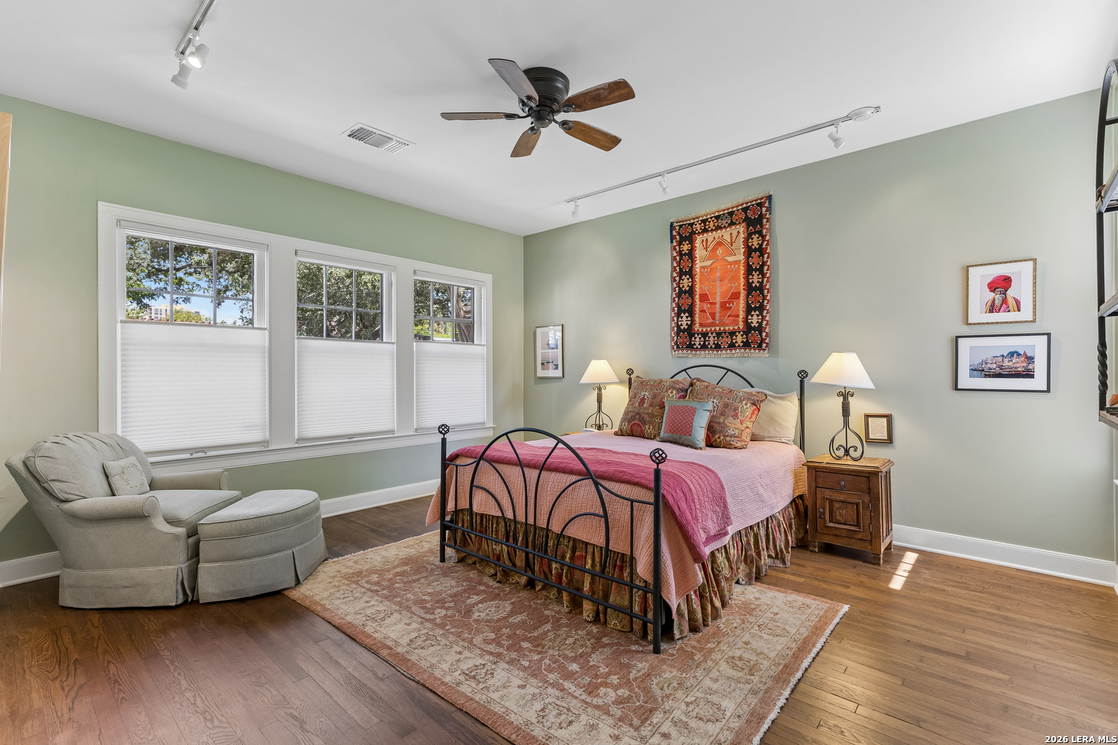 Bedroom with pink bedding, pale green walls, large shaded windows, and hardwood floors.