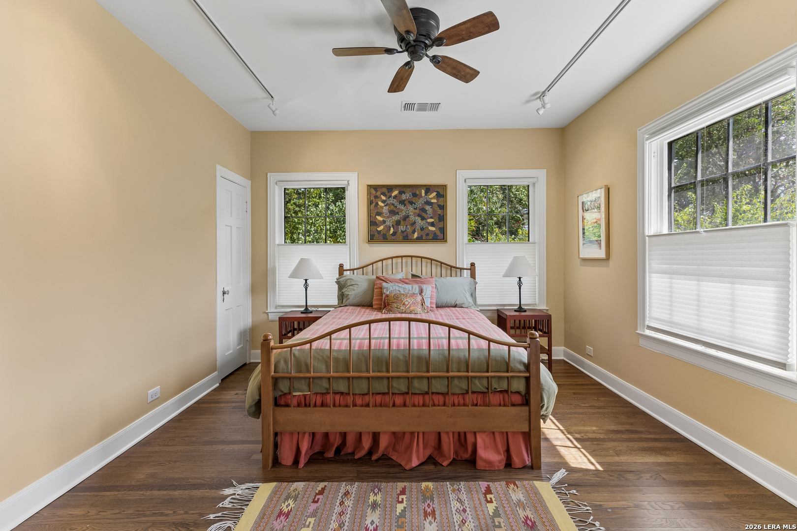 Bedroom with a wooden bed, pink and sage bedding, hardwood floors, and three windows.