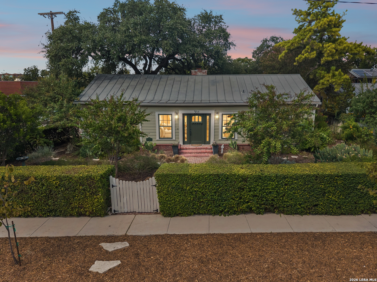 Single-story home with a gray metal roof, green front door, brick steps, and a hedge-lined garden.