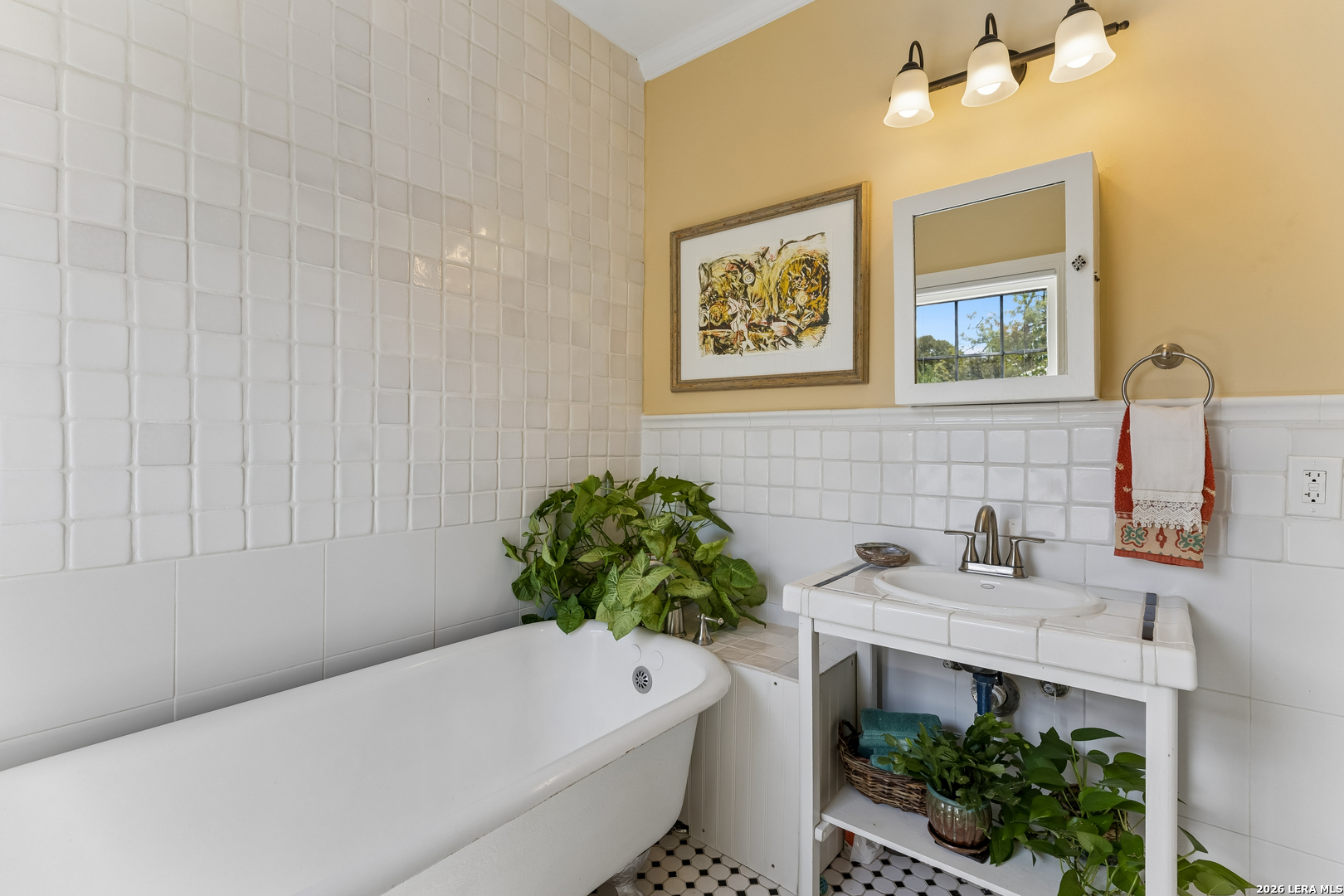 White-tiled bathroom with a bathtub, open-frame sink, leafy plants, and yellow walls.