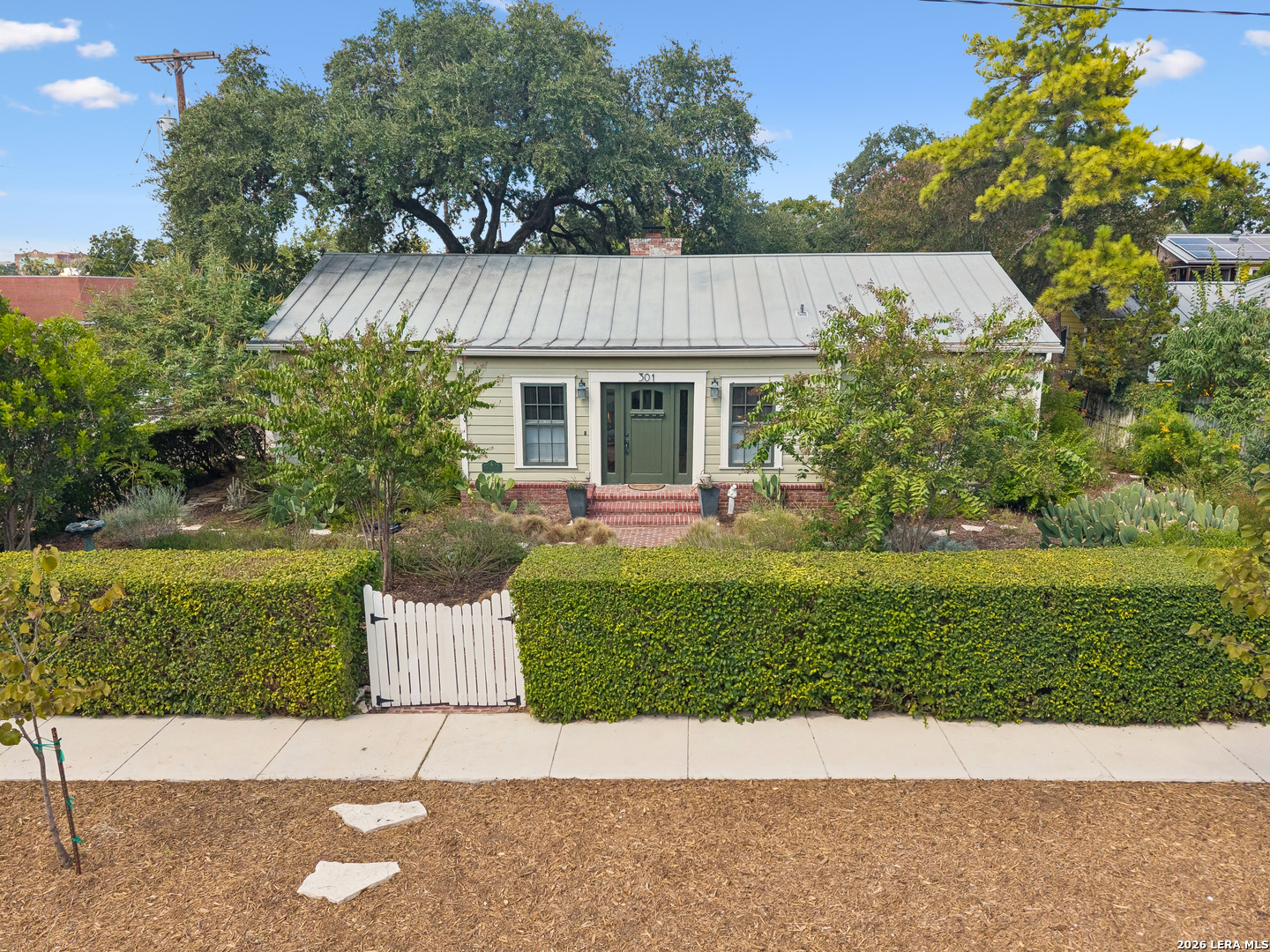 Single-story home with a metal roof, brick entry steps, and a white gate in the front hedge.