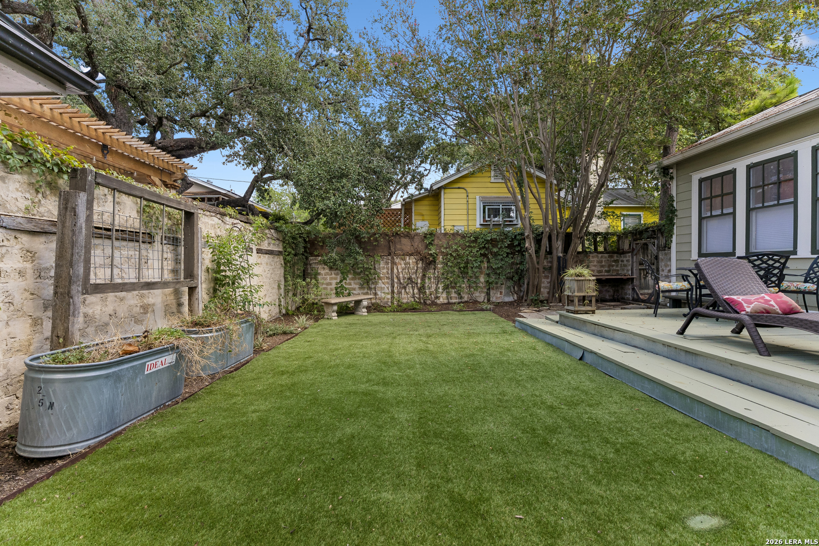 Backyard lawn with mature trees, a stone wall, and a raised deck with outdoor seating.