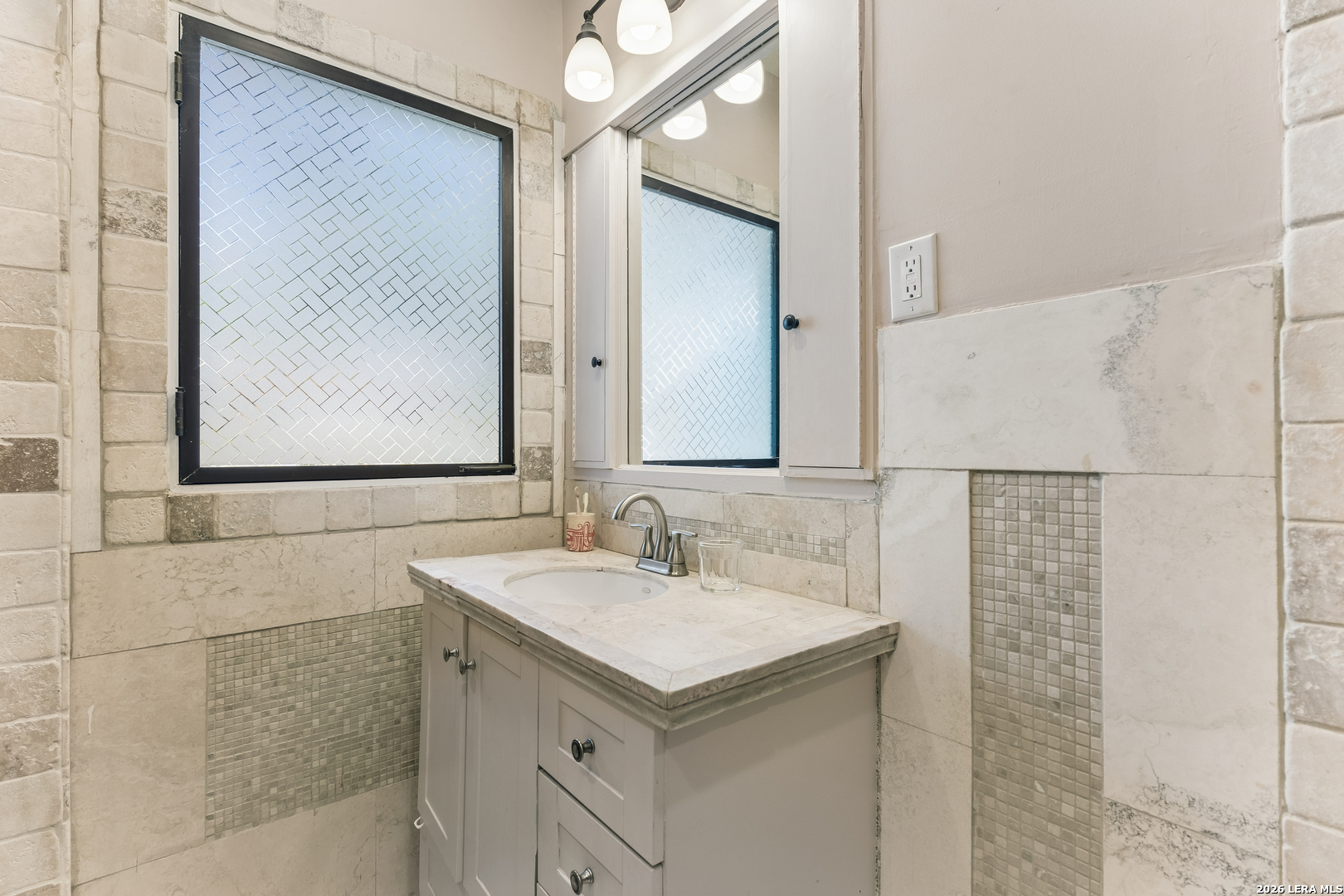 Bathroom vanity with a stone counter, frosted-glass windows, and marble-look tile accents.