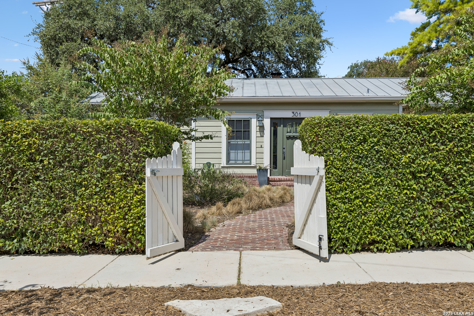 Single-story home behind an open white gate, with a brick entry path and tall hedges.