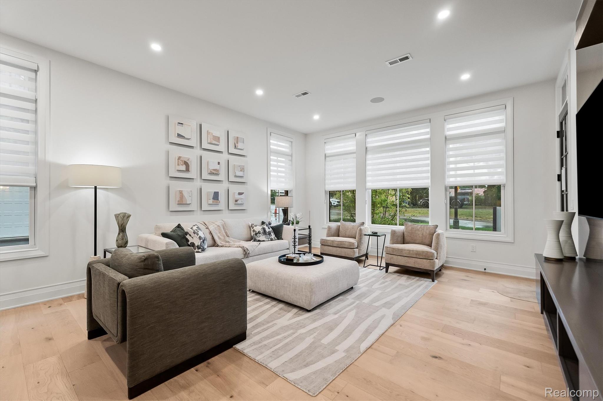 Living room with a sectional sofa, two armchairs, pale wood floors, and wide windows with striped shades.