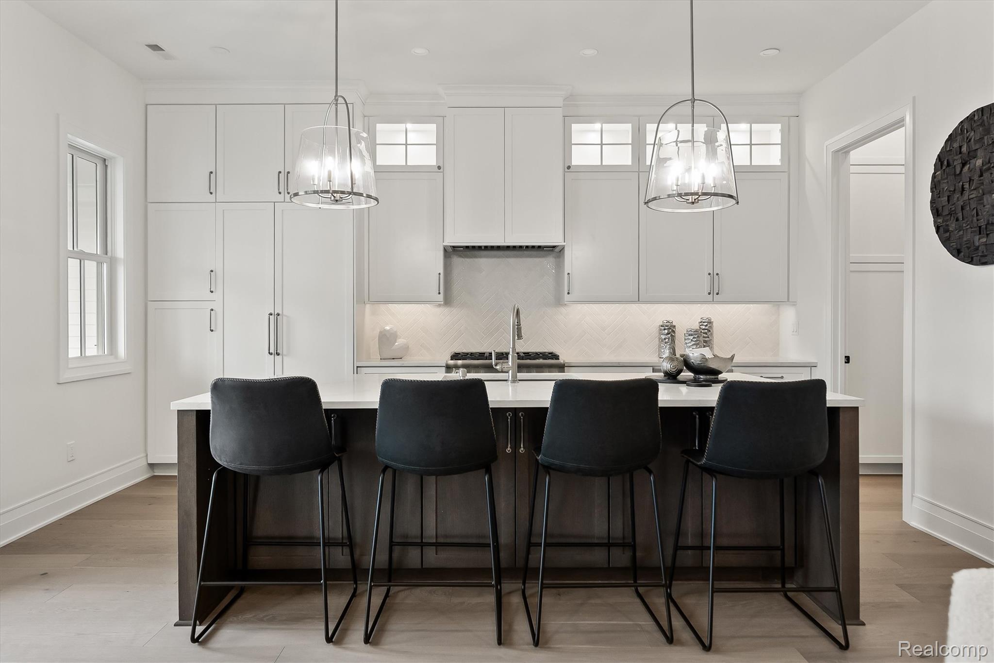 Modern white kitchen with dark island, four black stools, glass pendants, and herringbone backsplash.