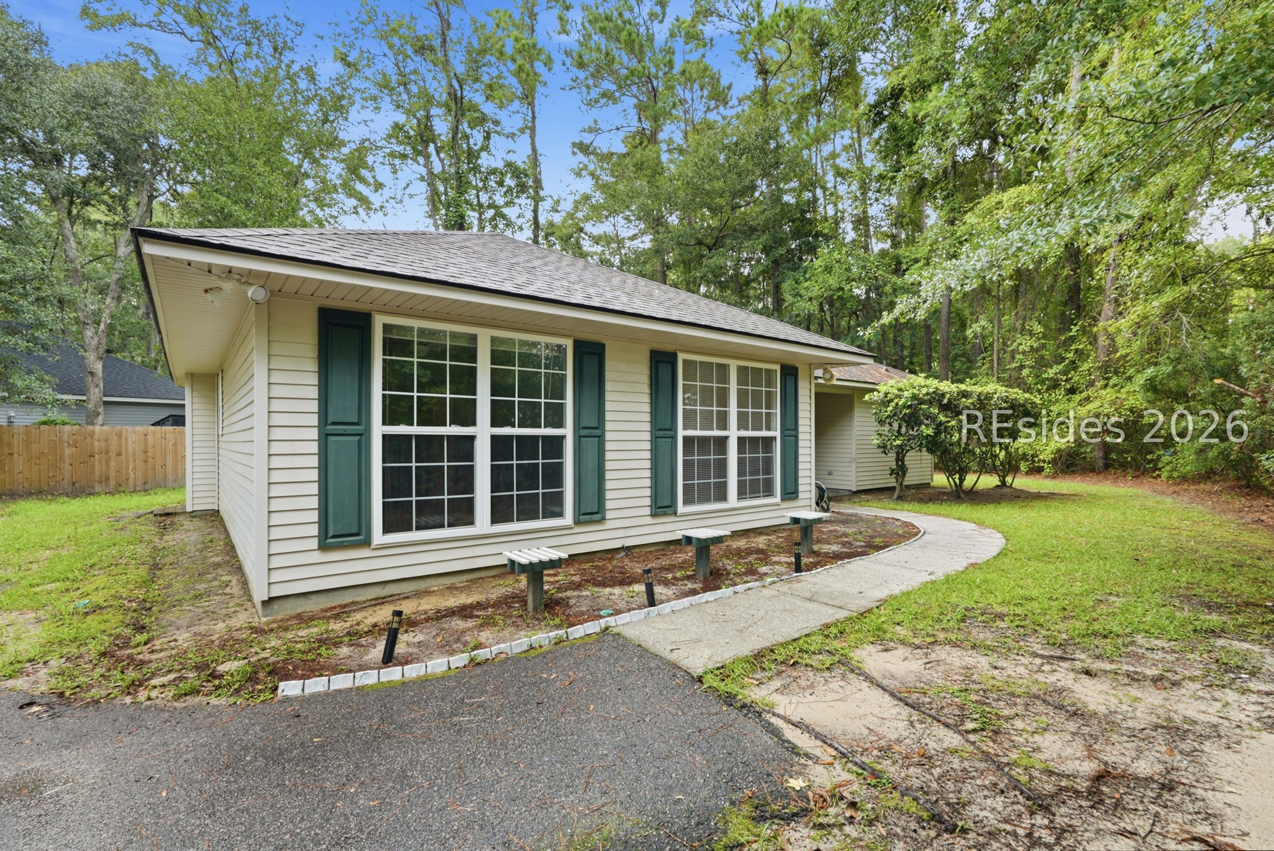 Single-story house with dark green shutters, a curved walkway, and mature trees surrounding the yard.
