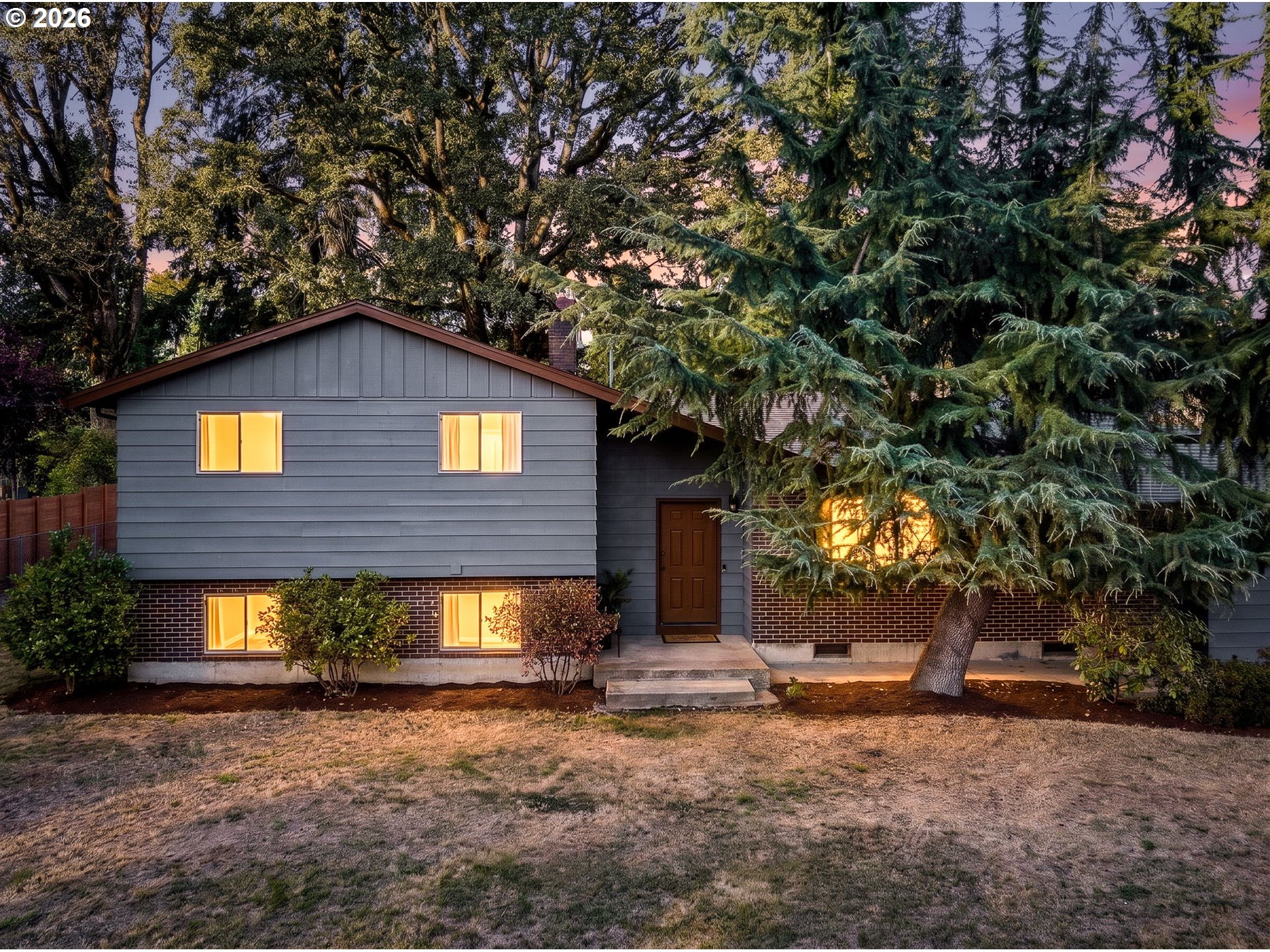 Gray split-level home with brick lower walls and glowing windows beneath mature trees at dusk.