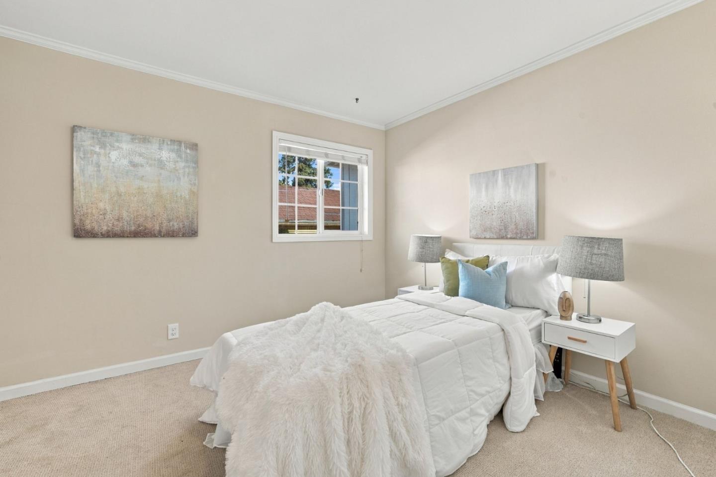Staged bedroom with a white bed, blue and green pillows, matching lamps, and a window beside it.