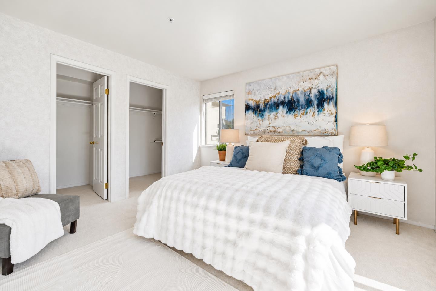 Staged bedroom with a white bed, blue pillows, abstract artwork, and two open closets.
