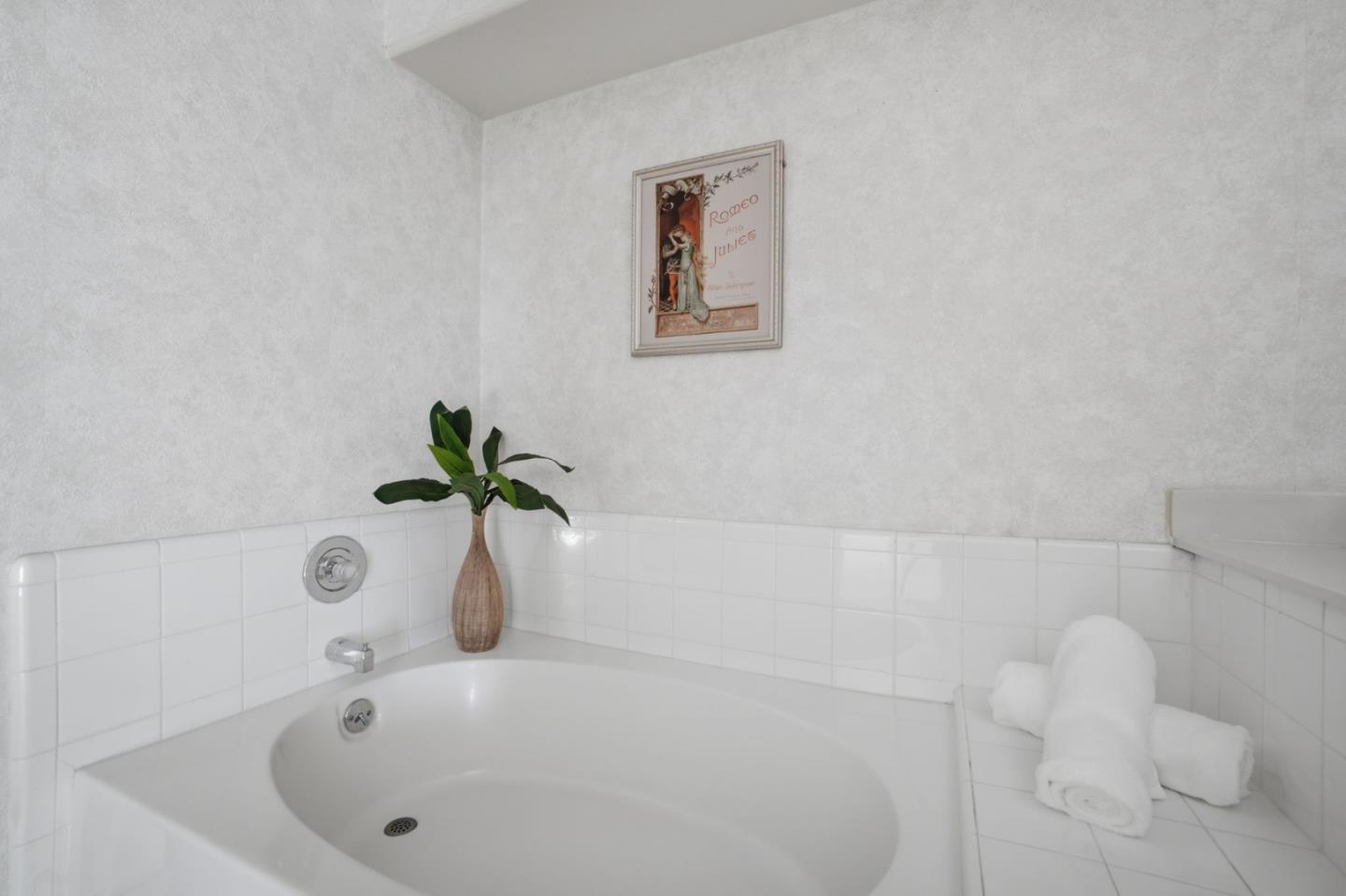 White soaking tub with tiled surround, rolled towels, and a potted plant.