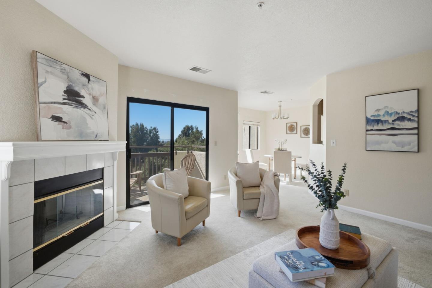 Living room with a tiled fireplace, cream armchairs, and sliding doors to a tree-lined balcony.