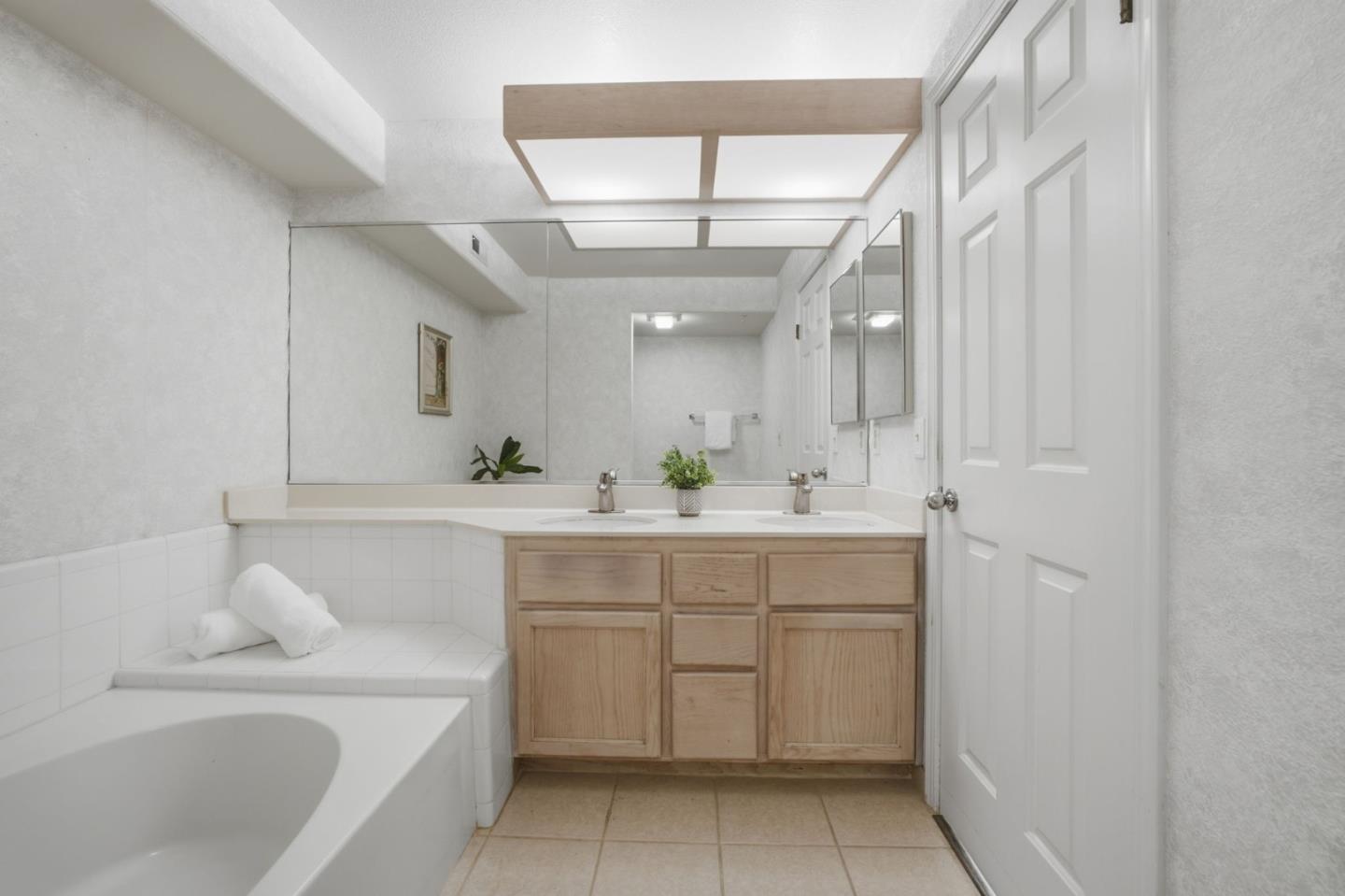 Bathroom with soaking tub, tiled surround, and light wood double vanity beneath a wall-length mirror.