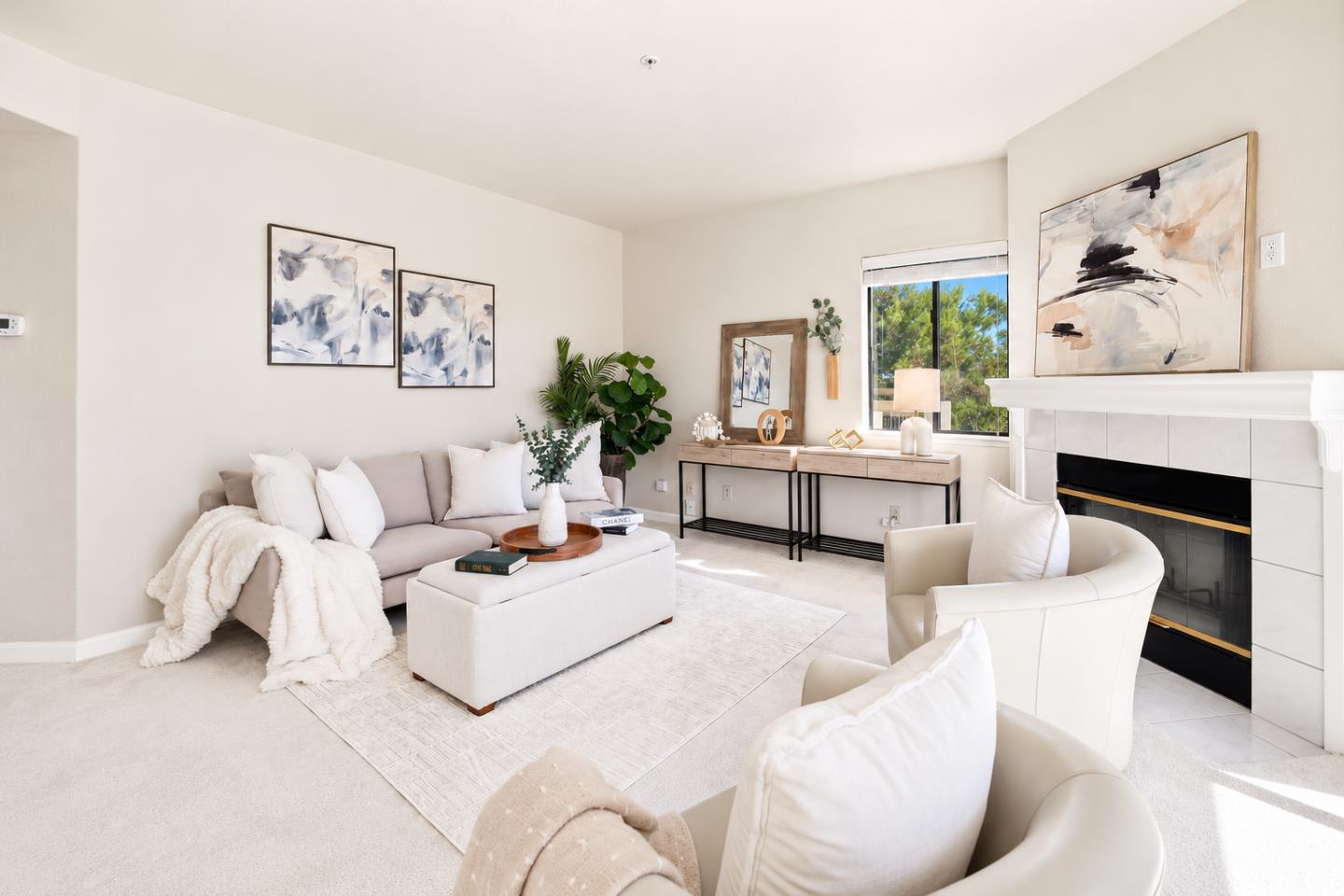 Staged living room with a white tiled fireplace, pale seating, abstract art, and a window overlooking greenery.