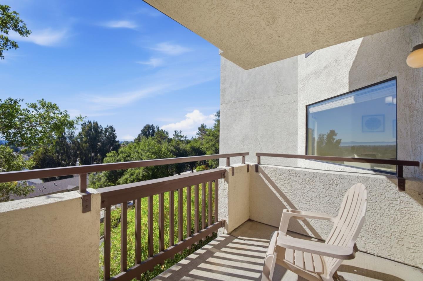 Covered balcony with a white Adirondack chair, brown railings, and a leafy view.