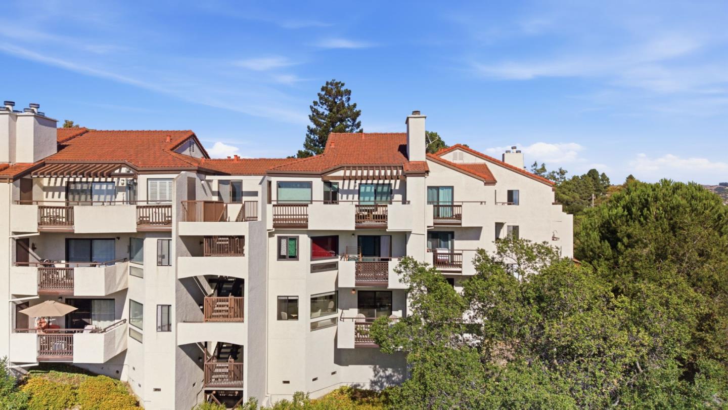 Condominium building with cream stucco walls, red tile roofs, and multiple balconies among mature trees.