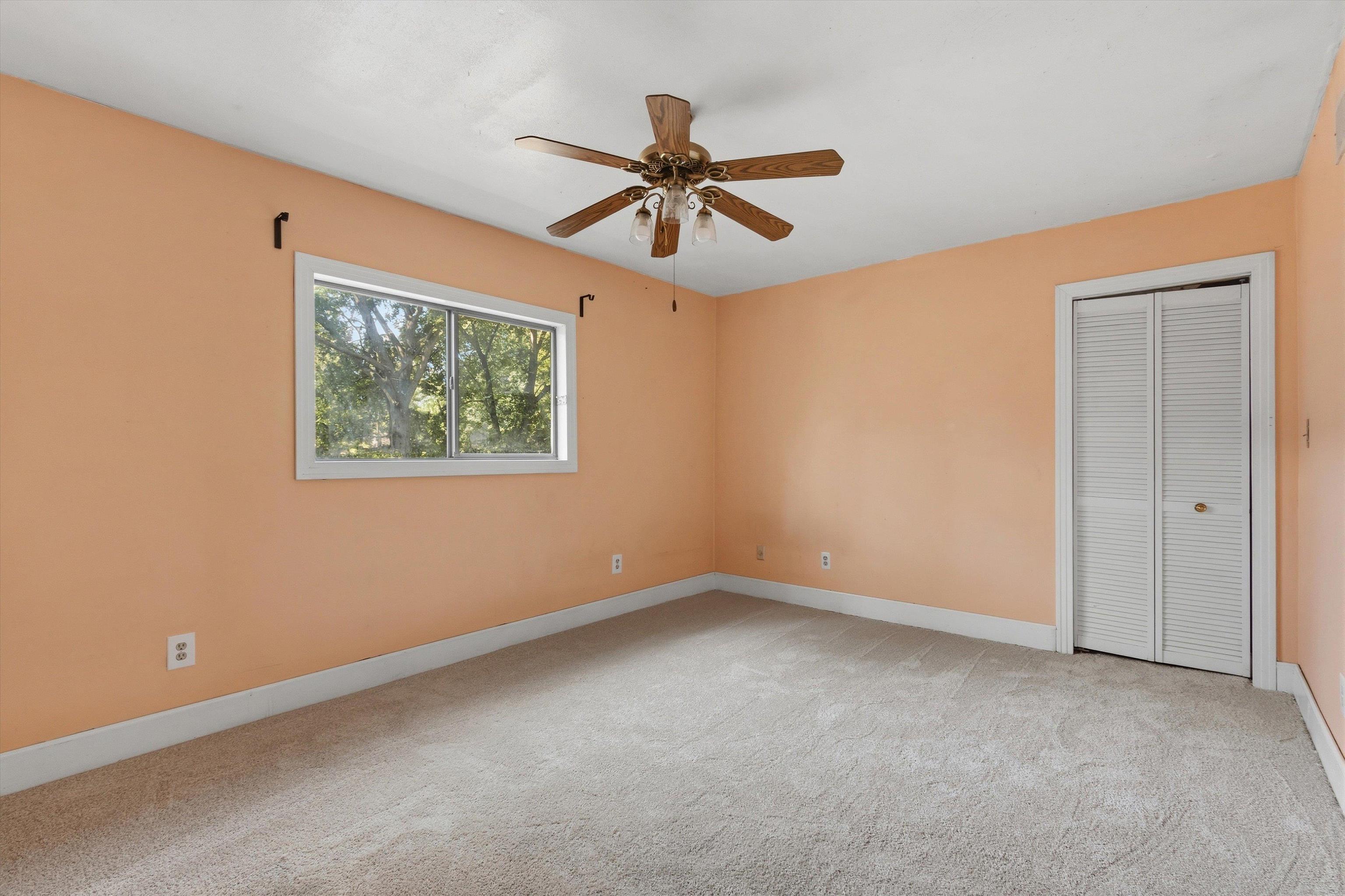 Empty bedroom with peach walls, beige carpet, a wood ceiling fan, and a white louvered closet.