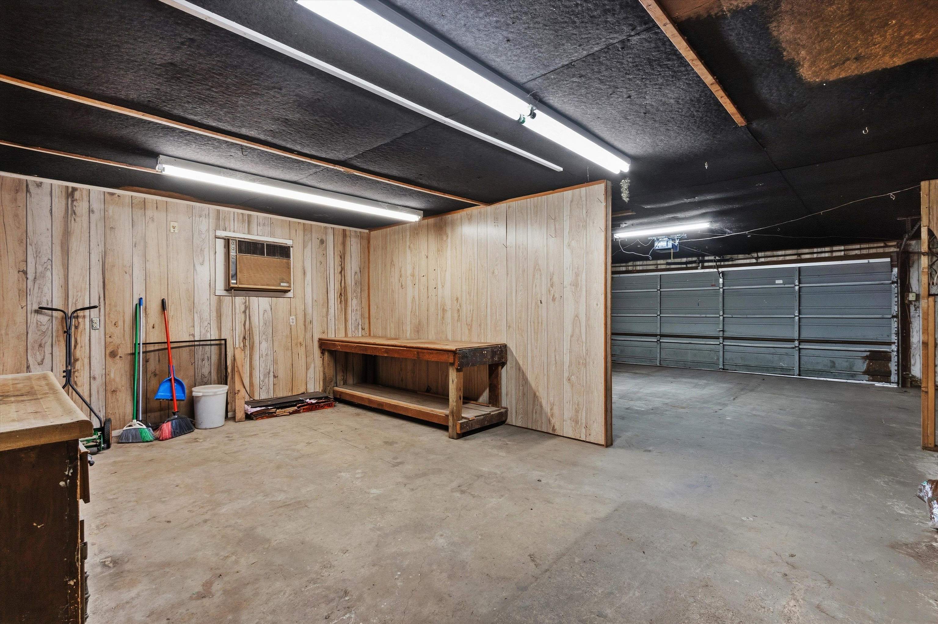 Garage workshop with a wooden workbench, cleaning tools, and a wide gray garage door.