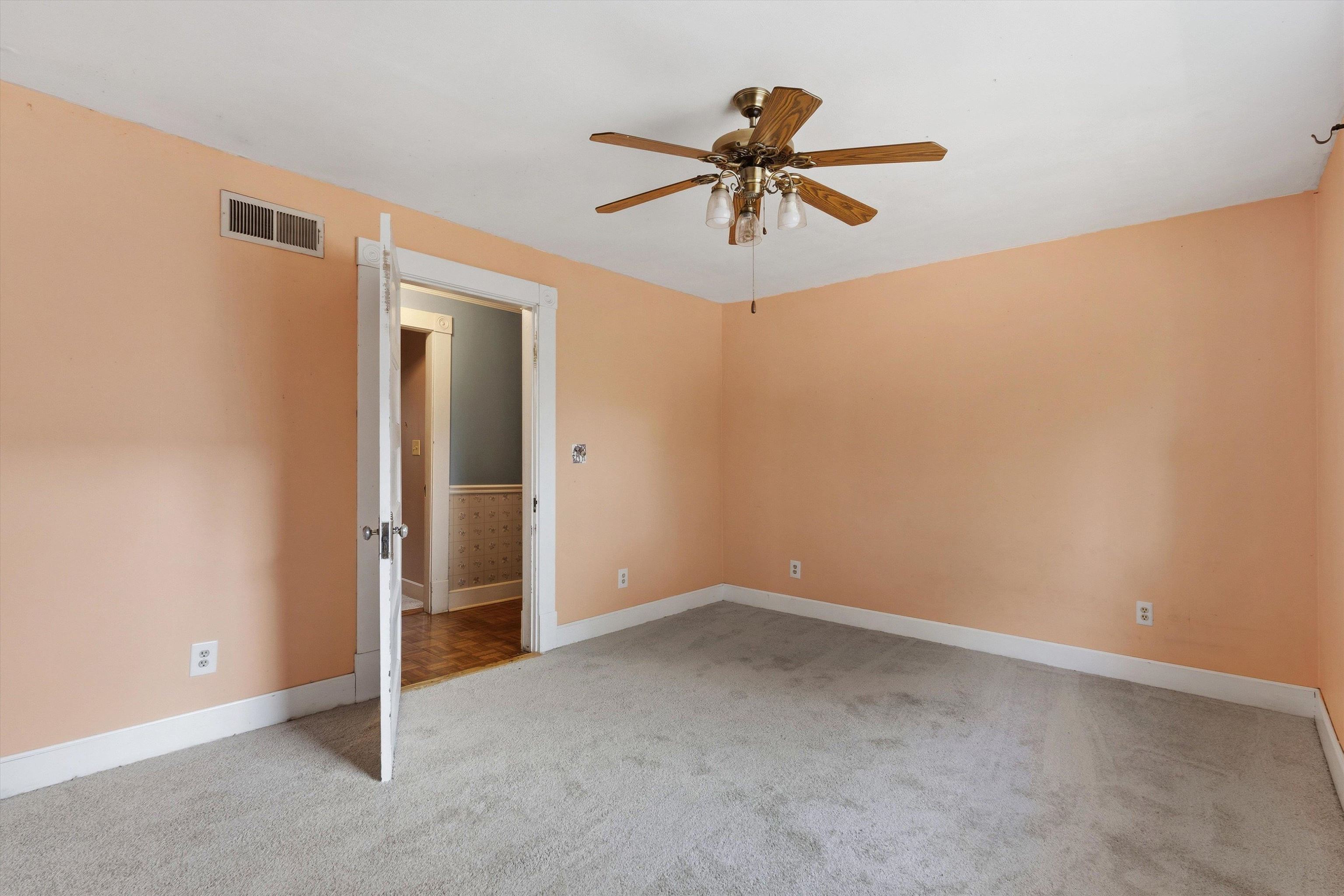 Empty peach-walled room with gray carpet, white trim, and a wood-bladed ceiling fan.