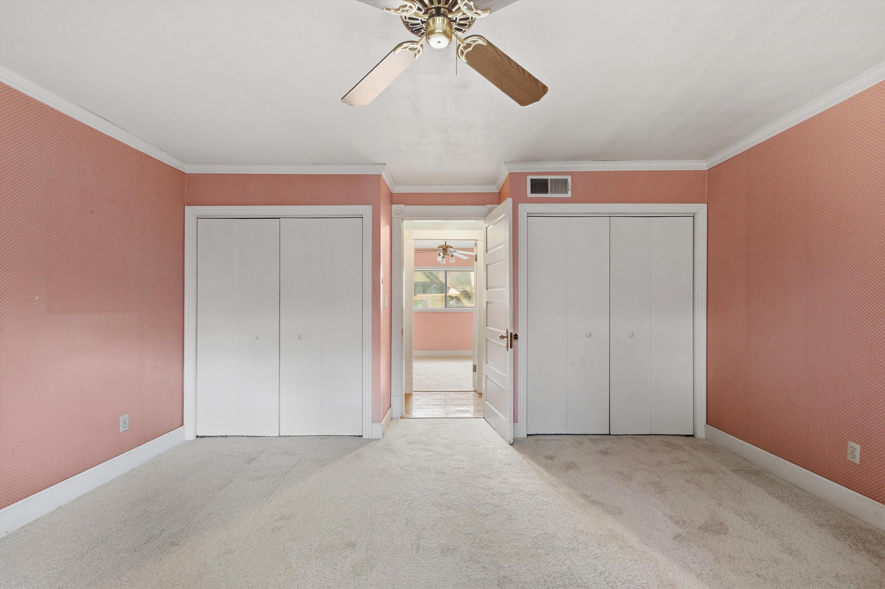 Empty bedroom with pink patterned walls, two white closets, and an open doorway to a sunlit room.