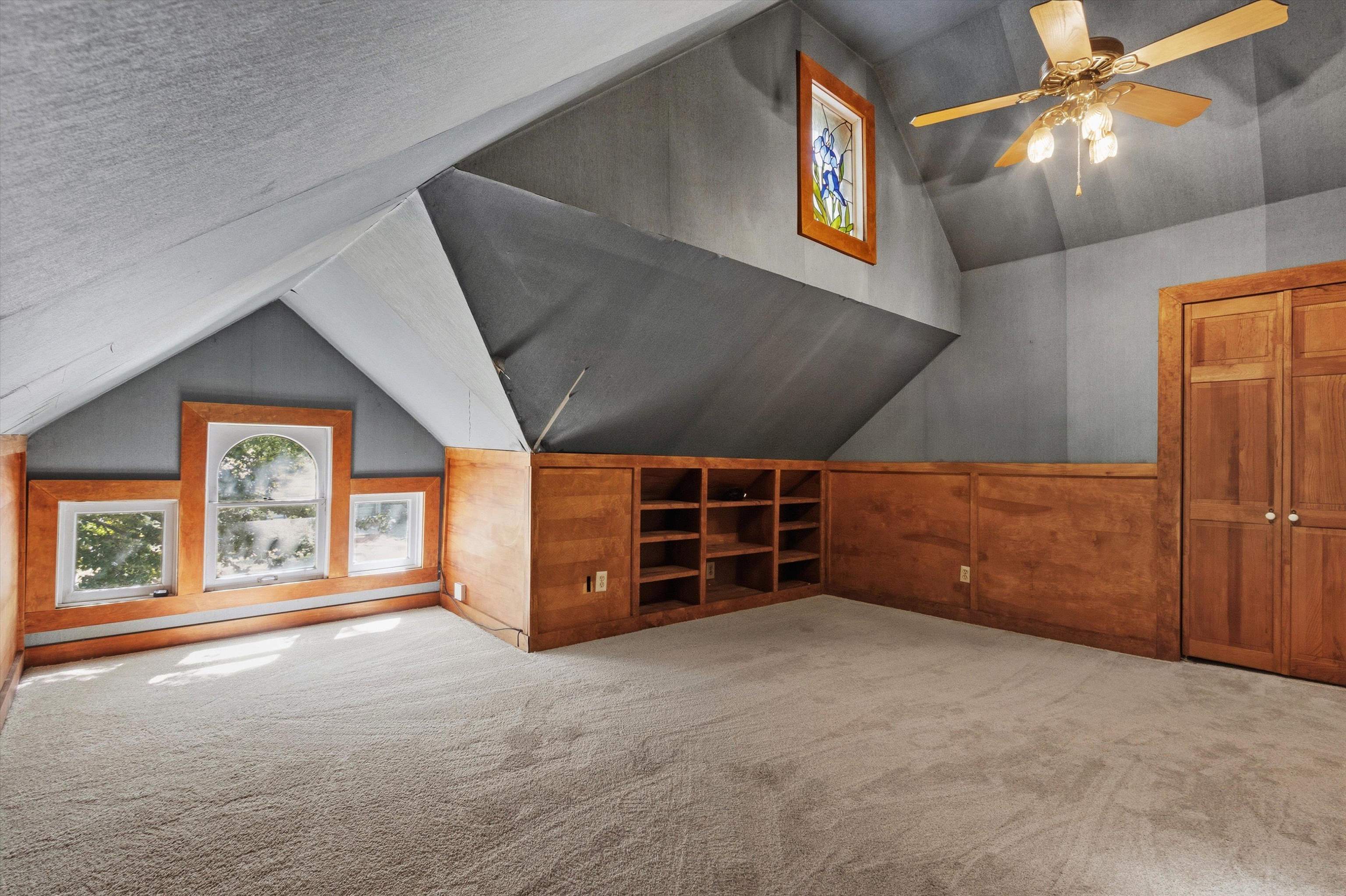 Attic room with a sloped ceiling, wood built-ins, low windows, and a ceiling fan.