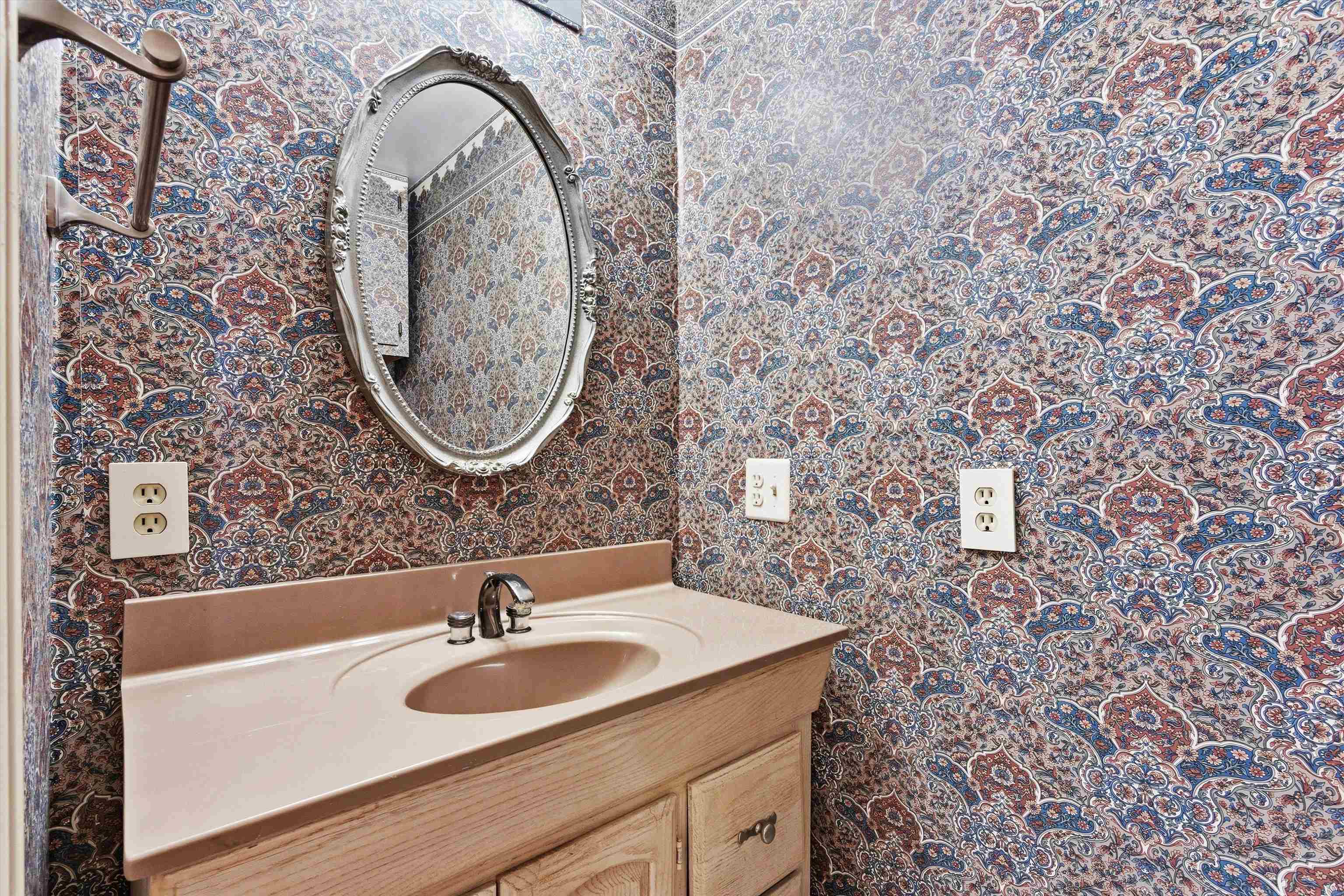 Bathroom vanity with an ornate oval mirror and densely patterned wallpaper.