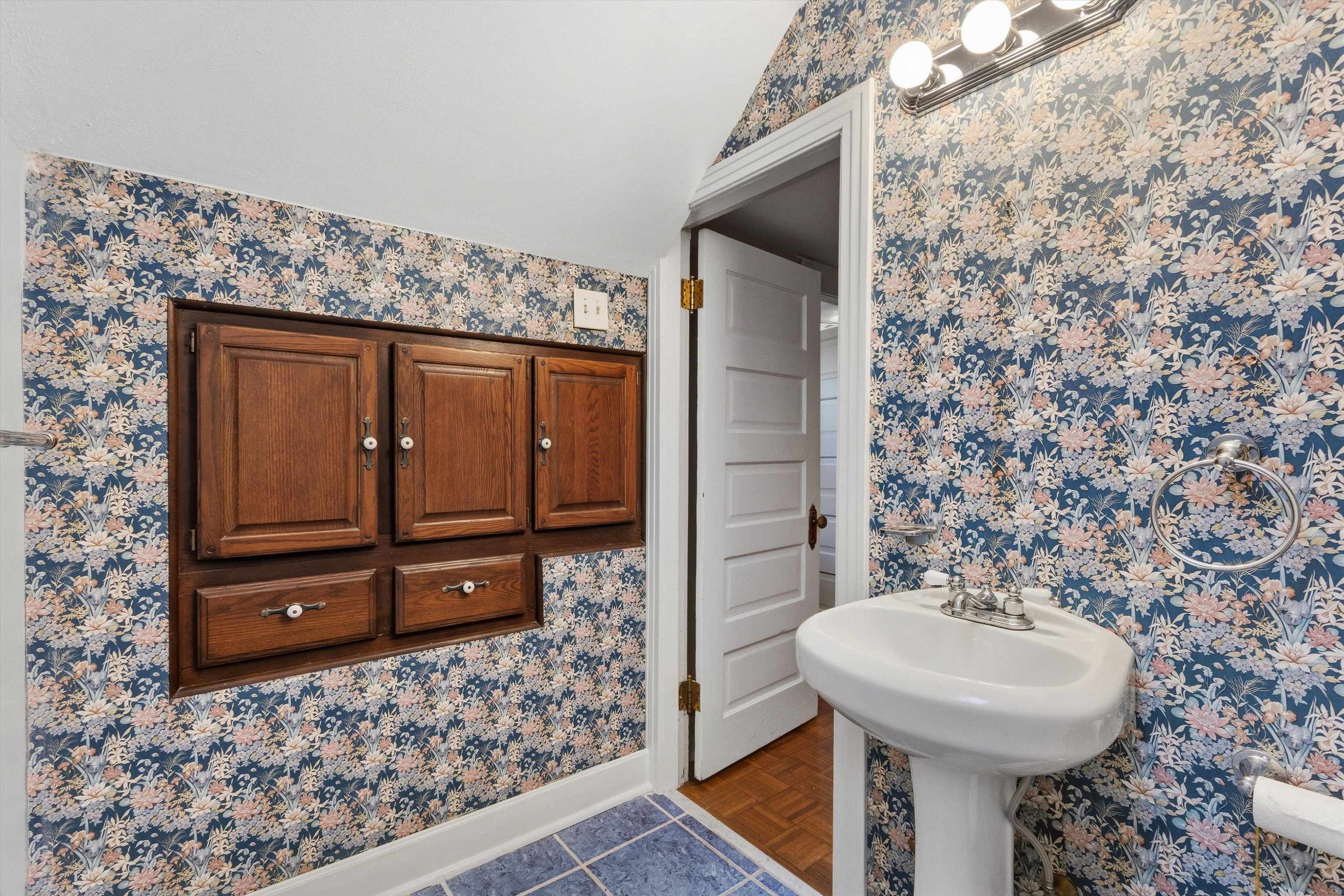 Bathroom with blue floral wallpaper, dark wood built-in cabinets, and a white pedestal sink.