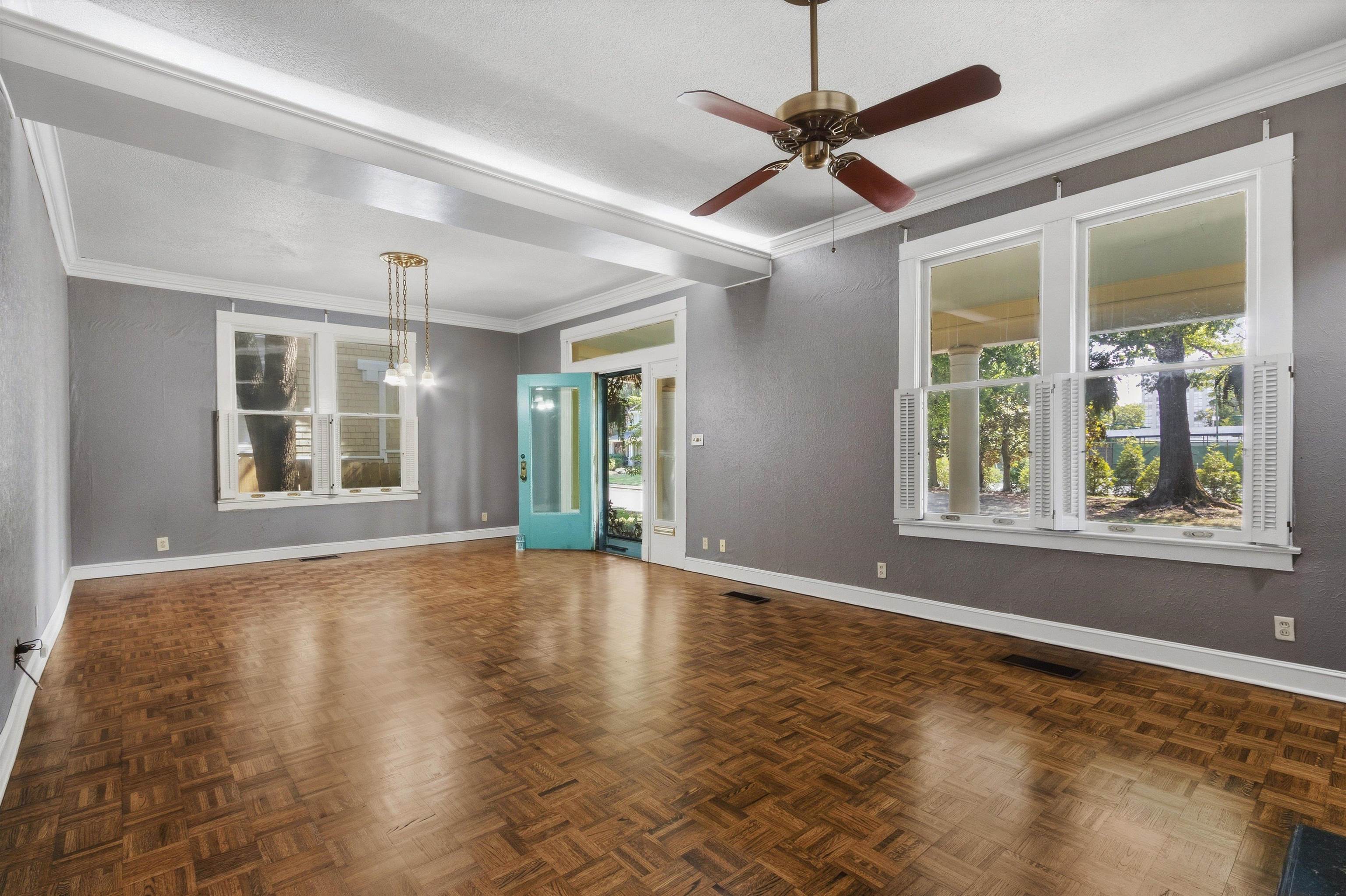 Empty living room with parquet floors, shuttered windows, and a turquoise front door.