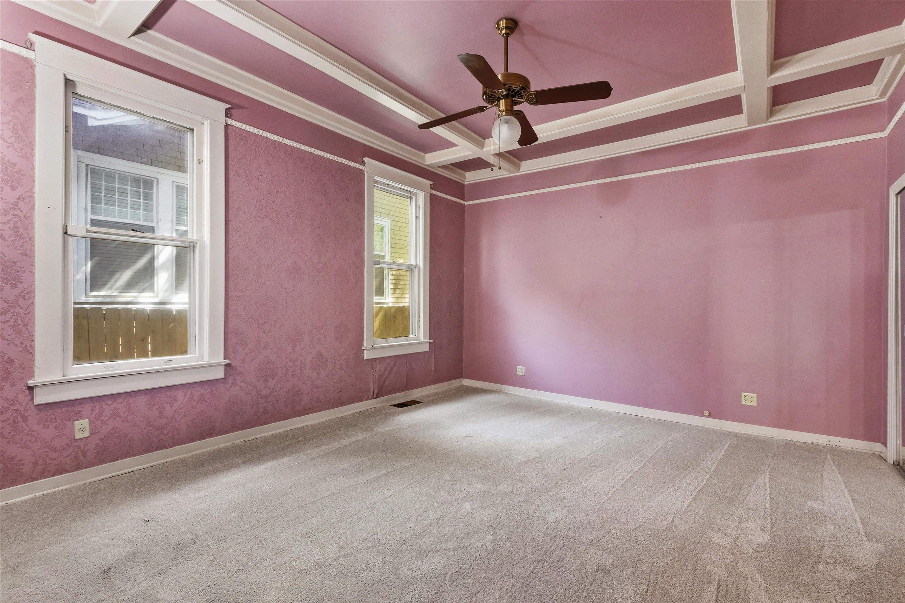 Empty bedroom with pink walls, a decorative coffered ceiling, ceiling fan, and two windows.