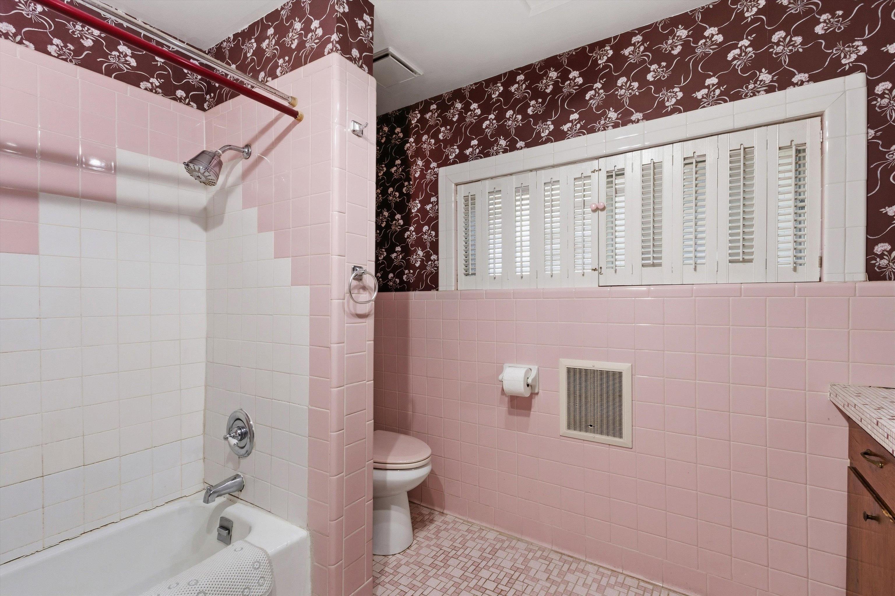 Bathroom with pink tile, a bathtub and shower, floral wallpaper, and a wide shuttered window.