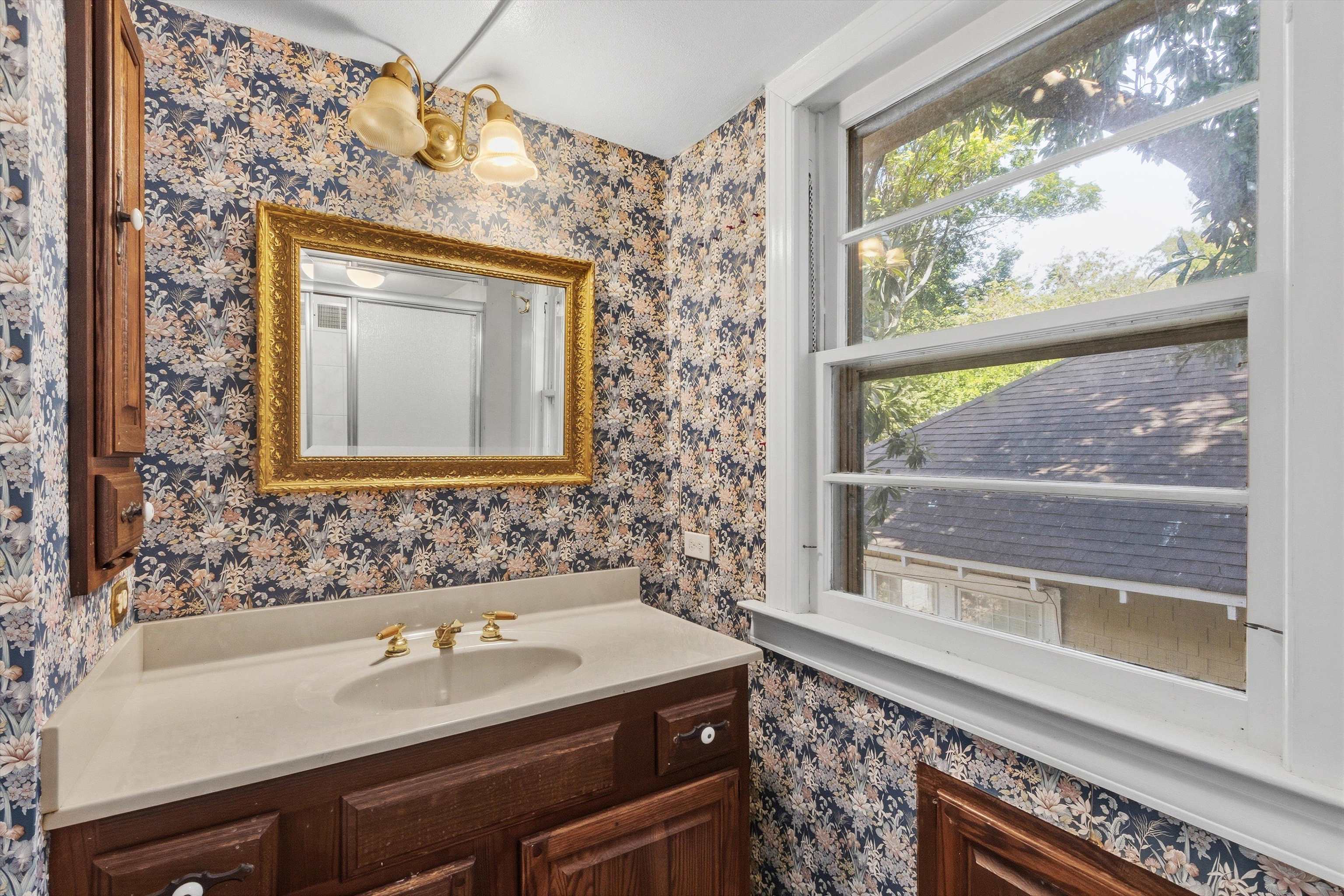 Bathroom vanity with floral wallpaper, an ornate gold mirror, and a large window.