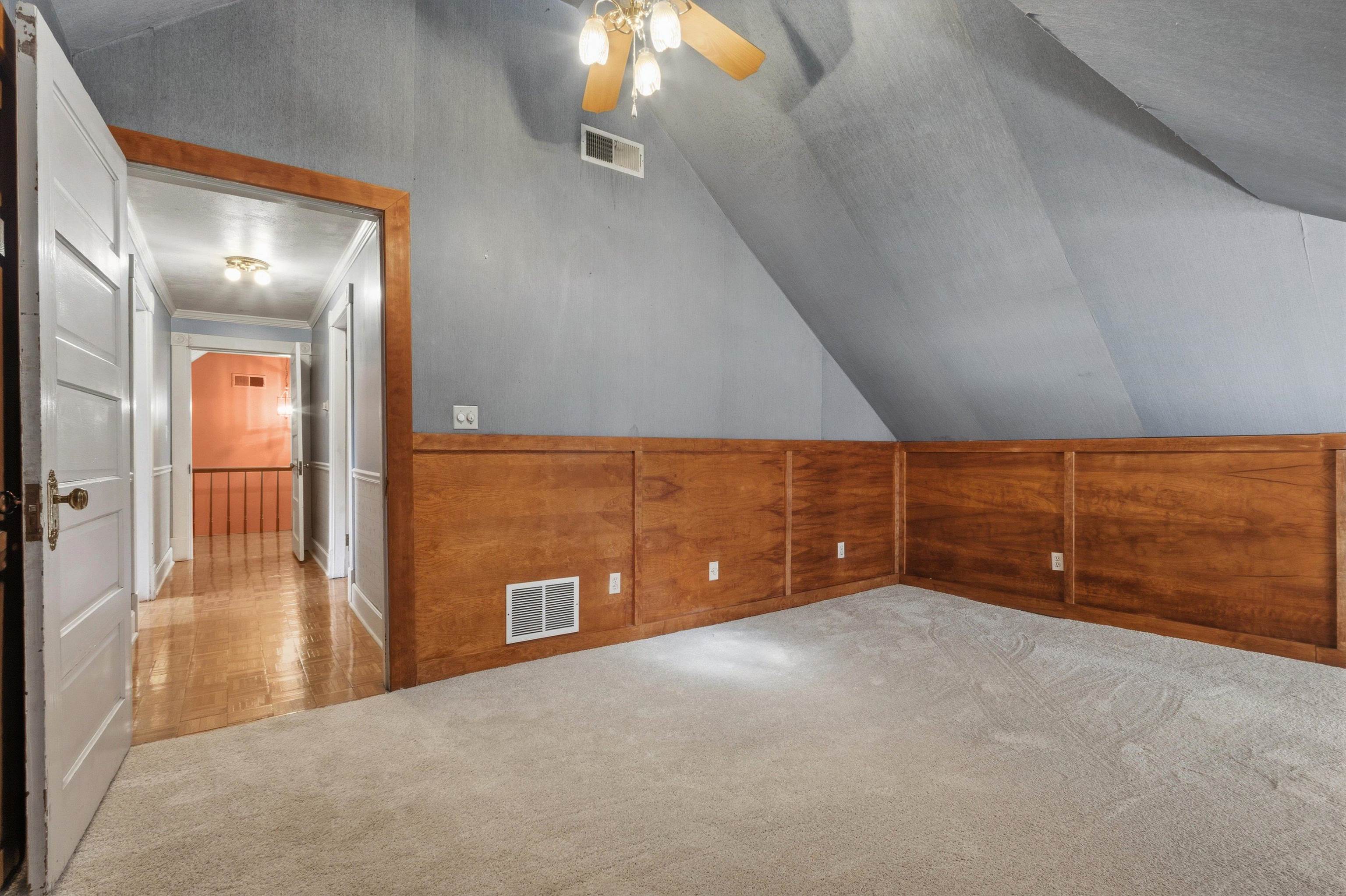 Finished attic room with sloped ceiling, wood wainscoting, beige carpet, and an open doorway to the hall.