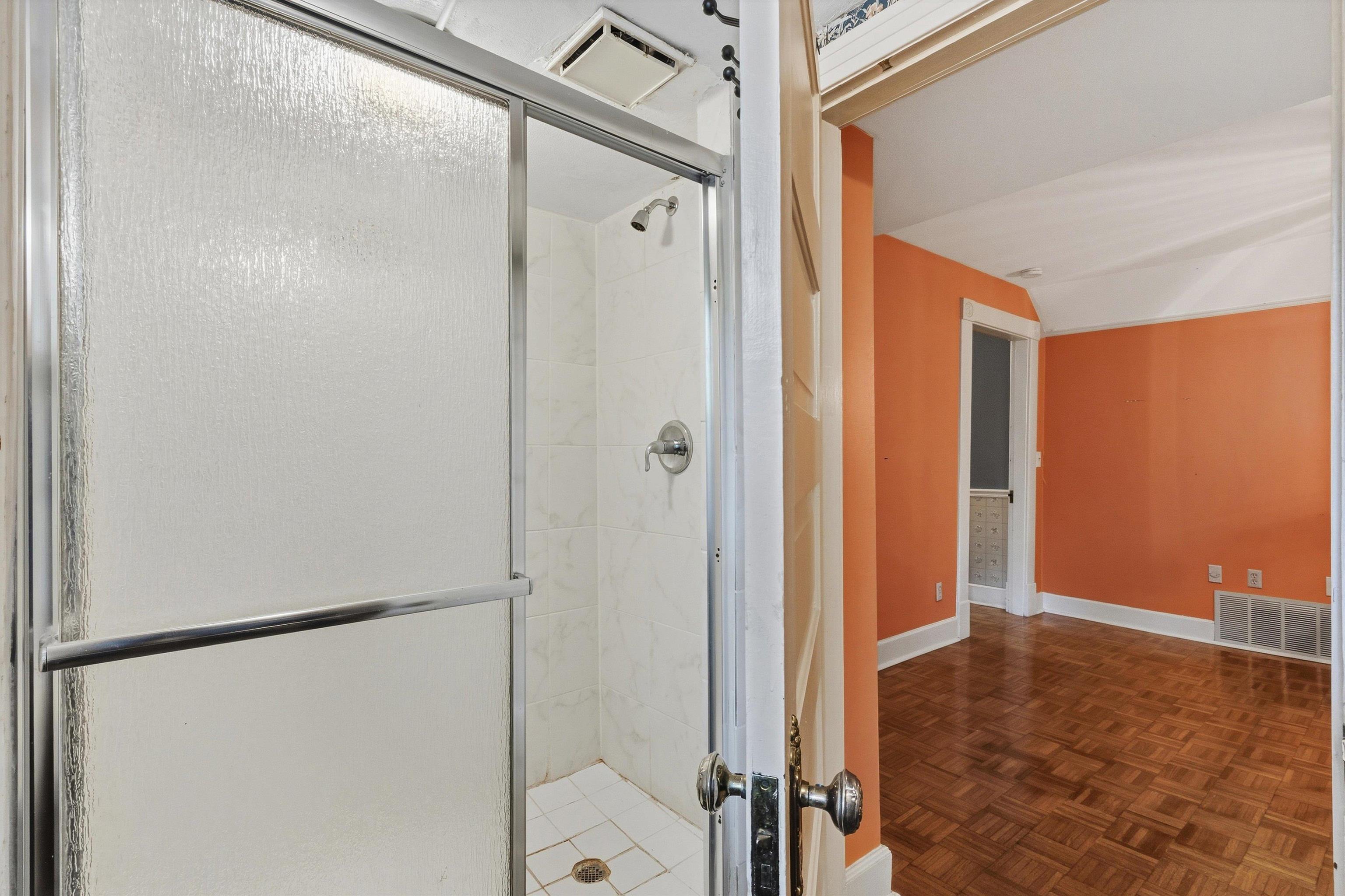 Tiled shower stall beside an orange room with parquet flooring.