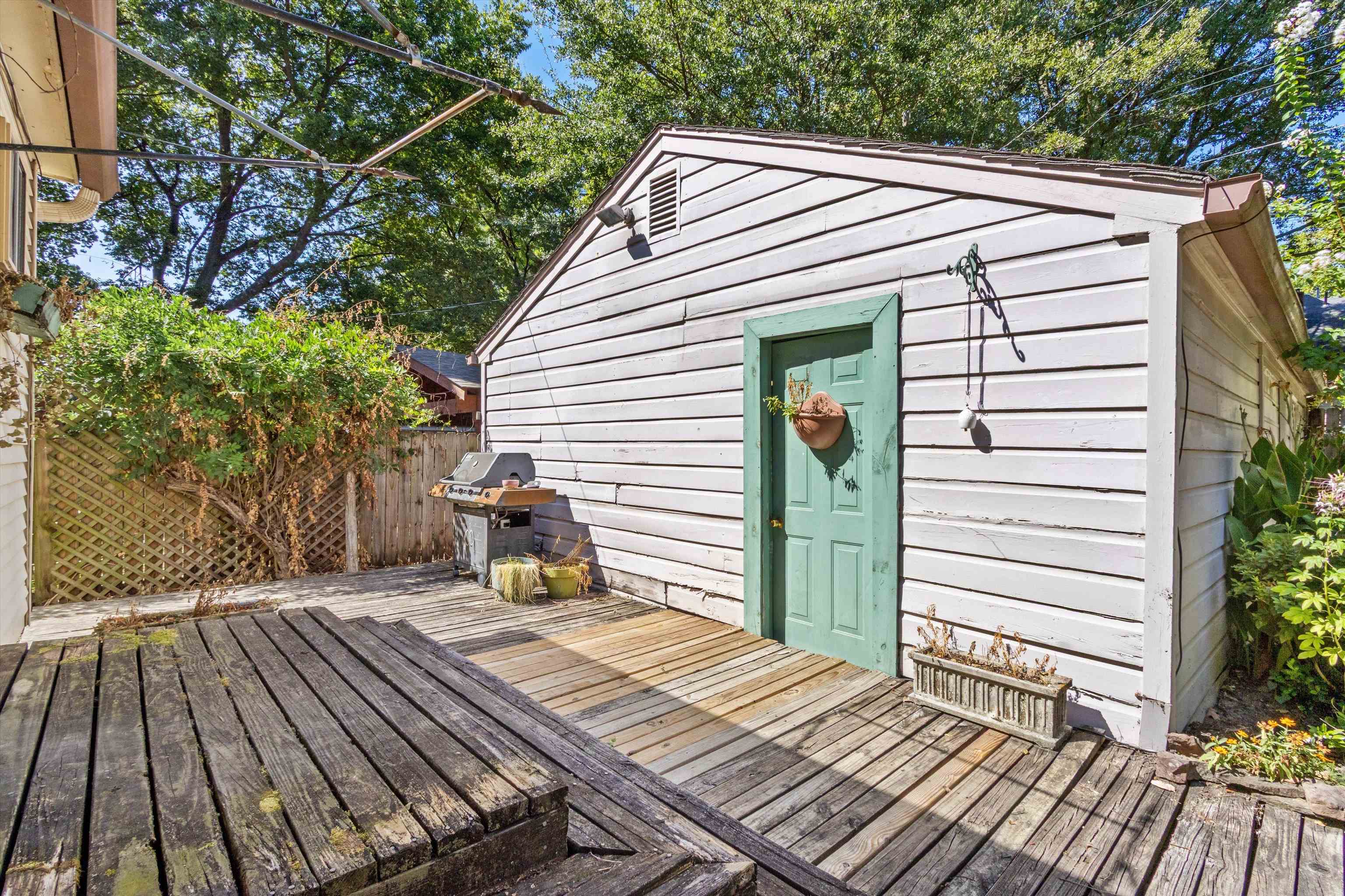 Backyard deck beside a white outbuilding with a green door, grill, and lattice fence.