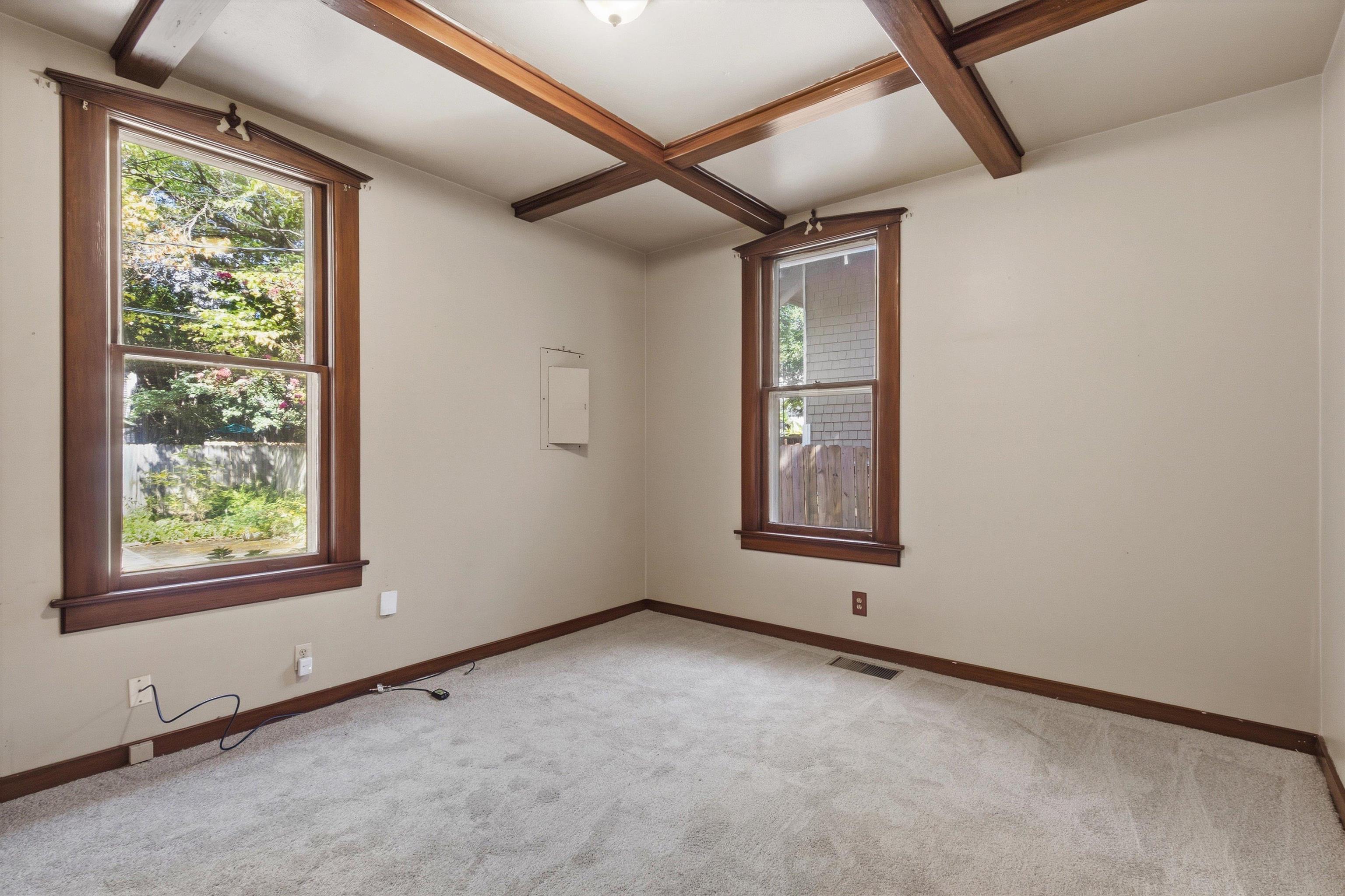 Empty room with dark wood ceiling beams, two tall windows, and light carpet.