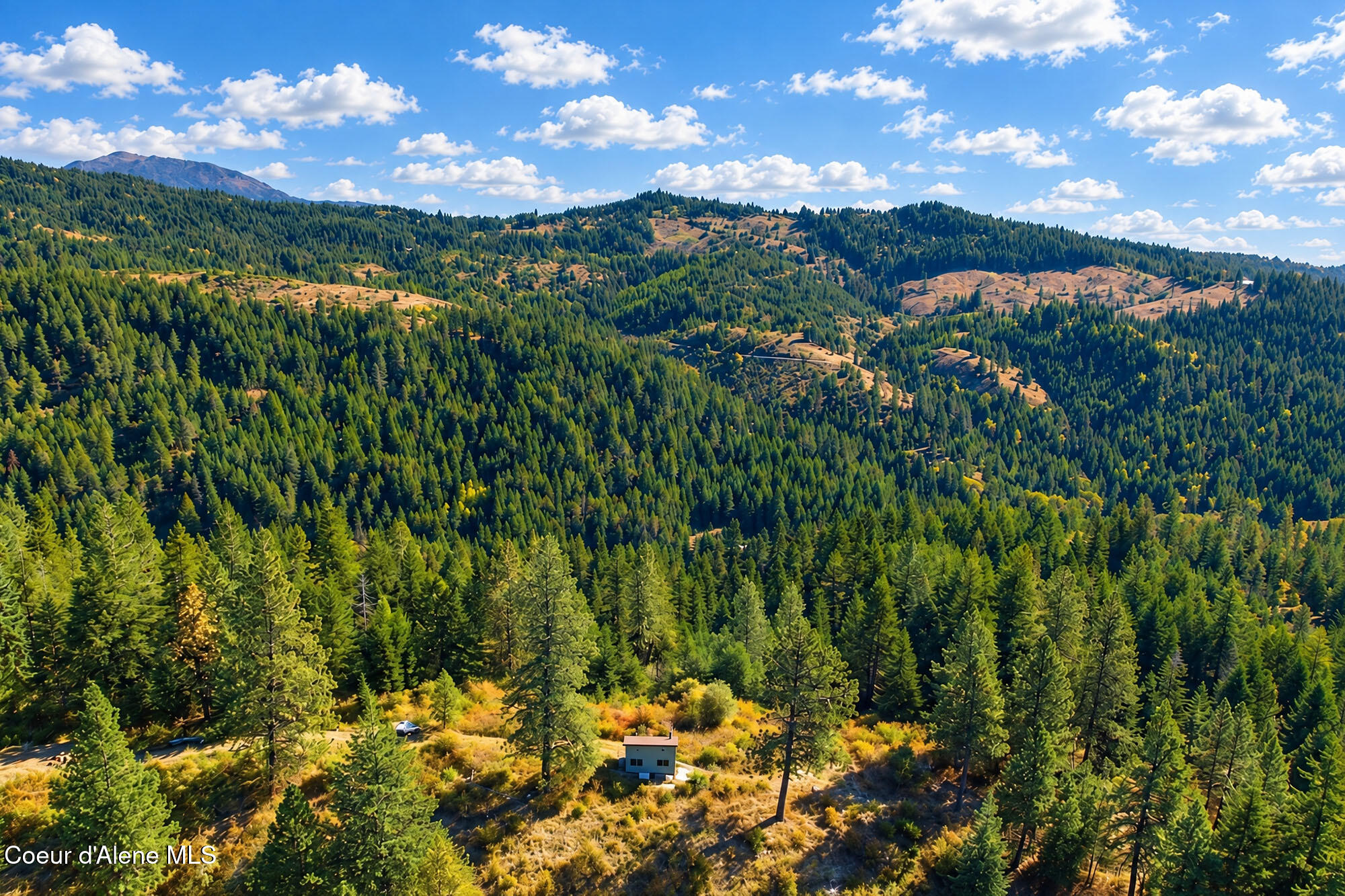 Aerial view of a small building in a forest clearing, with rolling wooded hills and distant mountains.