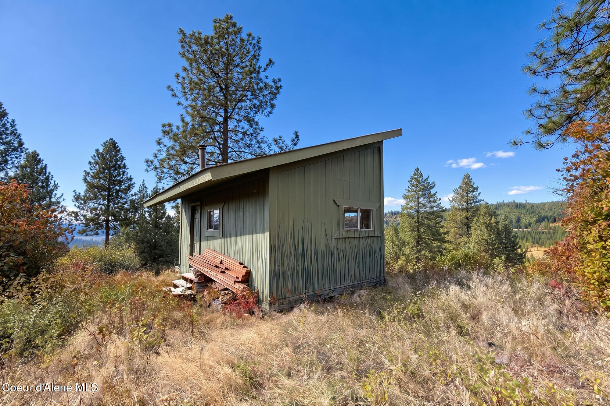 Small green cabin among pine trees, with stacked lumber beside it and wooded hills beyond.
