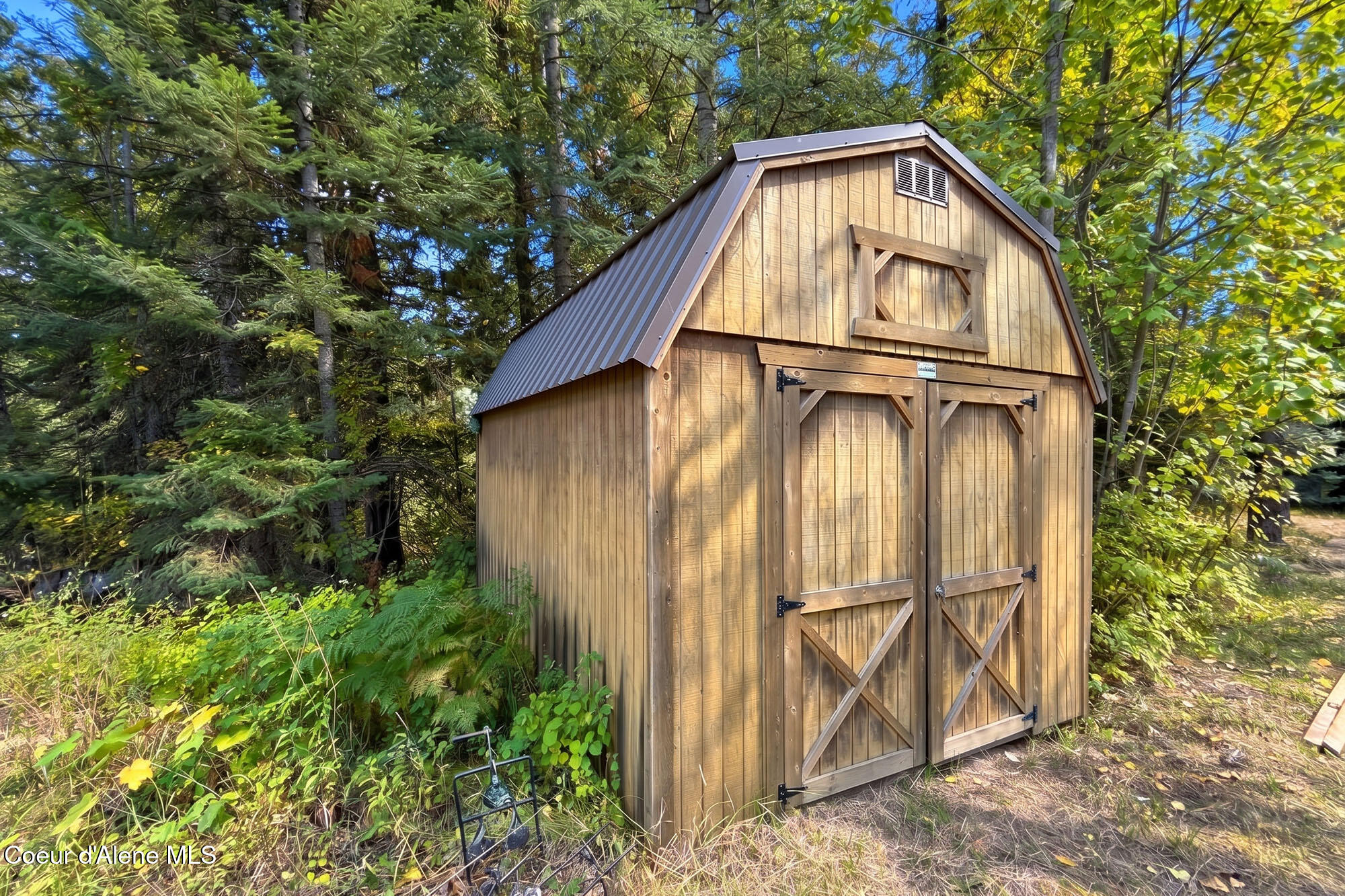 Wooden barn-style storage shed with a metal roof among trees and ferns.