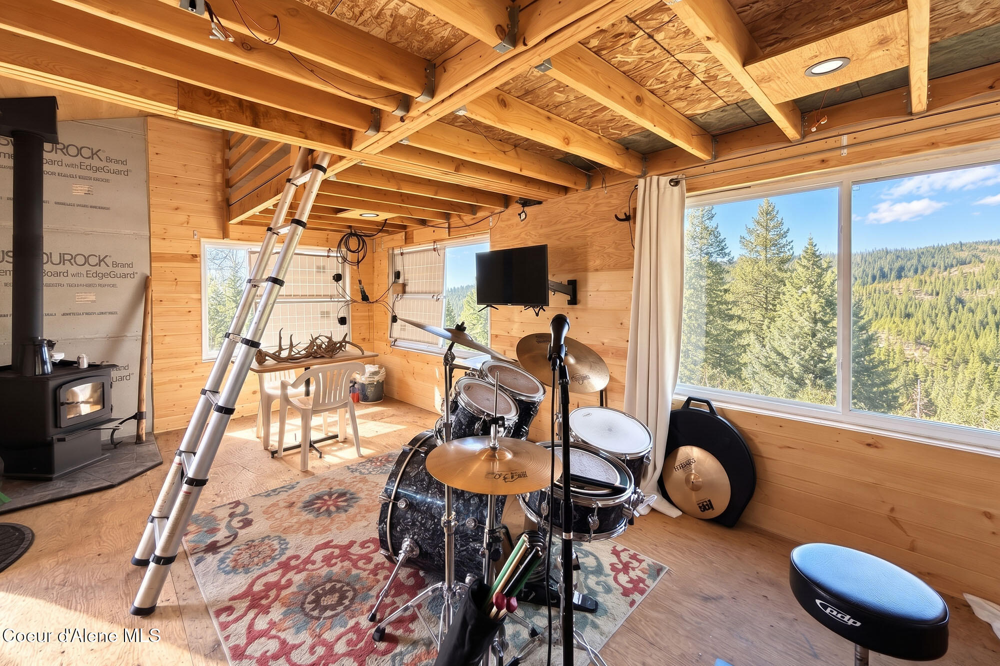 Wood-paneled music room with a drum kit, wood stove, and large windows overlooking a forested hillside.