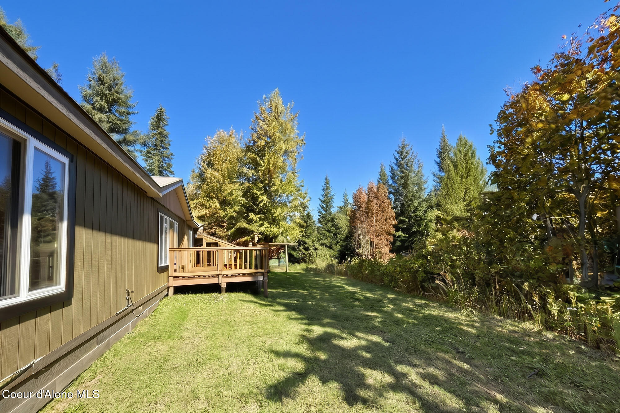 Brown house and wooden deck beside a grassy yard bordered by autumn trees and evergreens.