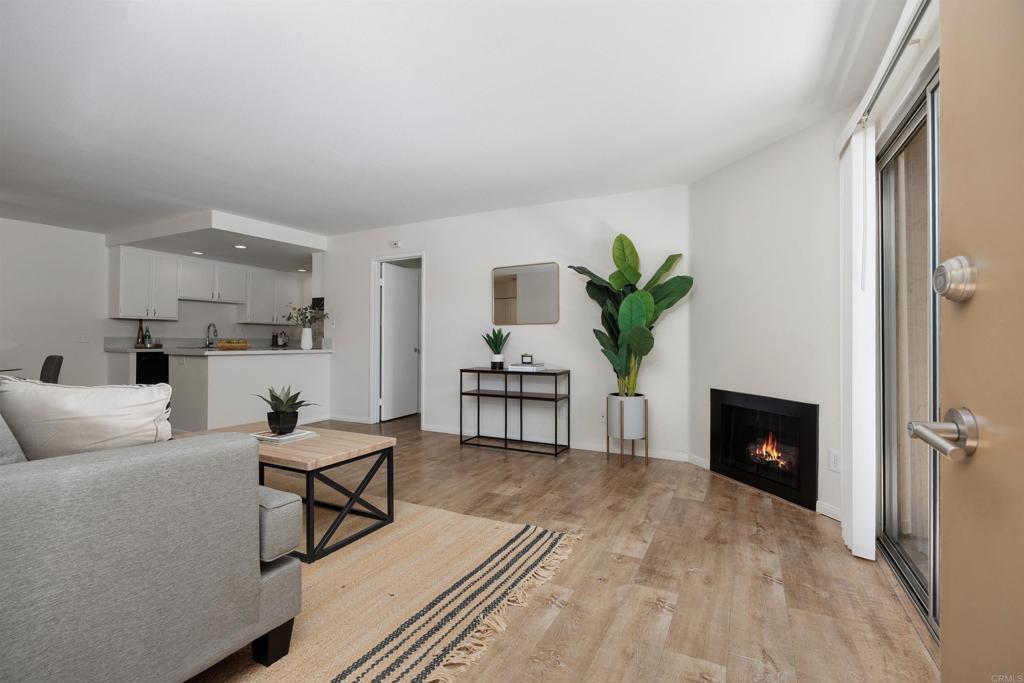 Open-plan living room with a gray sofa, wood floors, fireplace, and adjoining white kitchen.